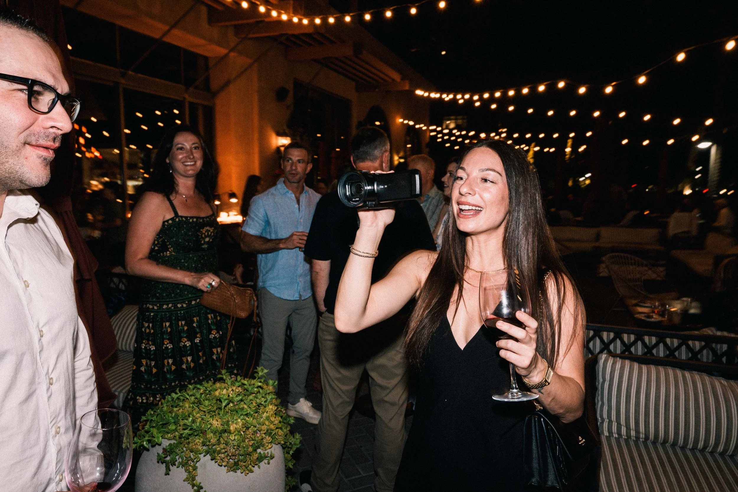 Bridesmaid films at wedding with camcorder at night with wine glass in hand and cute hanging lights in background