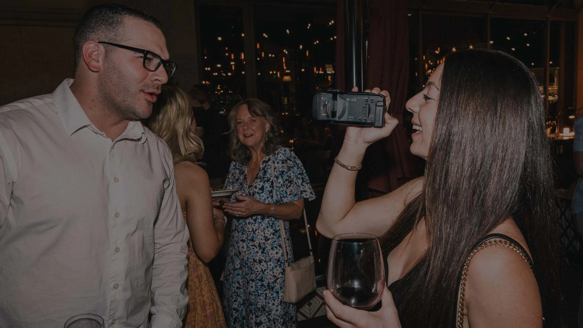 A guest films a candid moment with a camcorder rental at an indoor evening celebration, holding a glass of red wine as warm city lights glow through the windows behind the group.