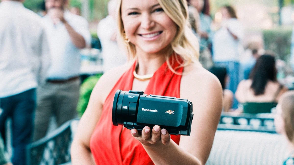 A woman in a red dresses holds a panasonic 4k camcorder in front of her, presenting it, she is at a wedding reception.