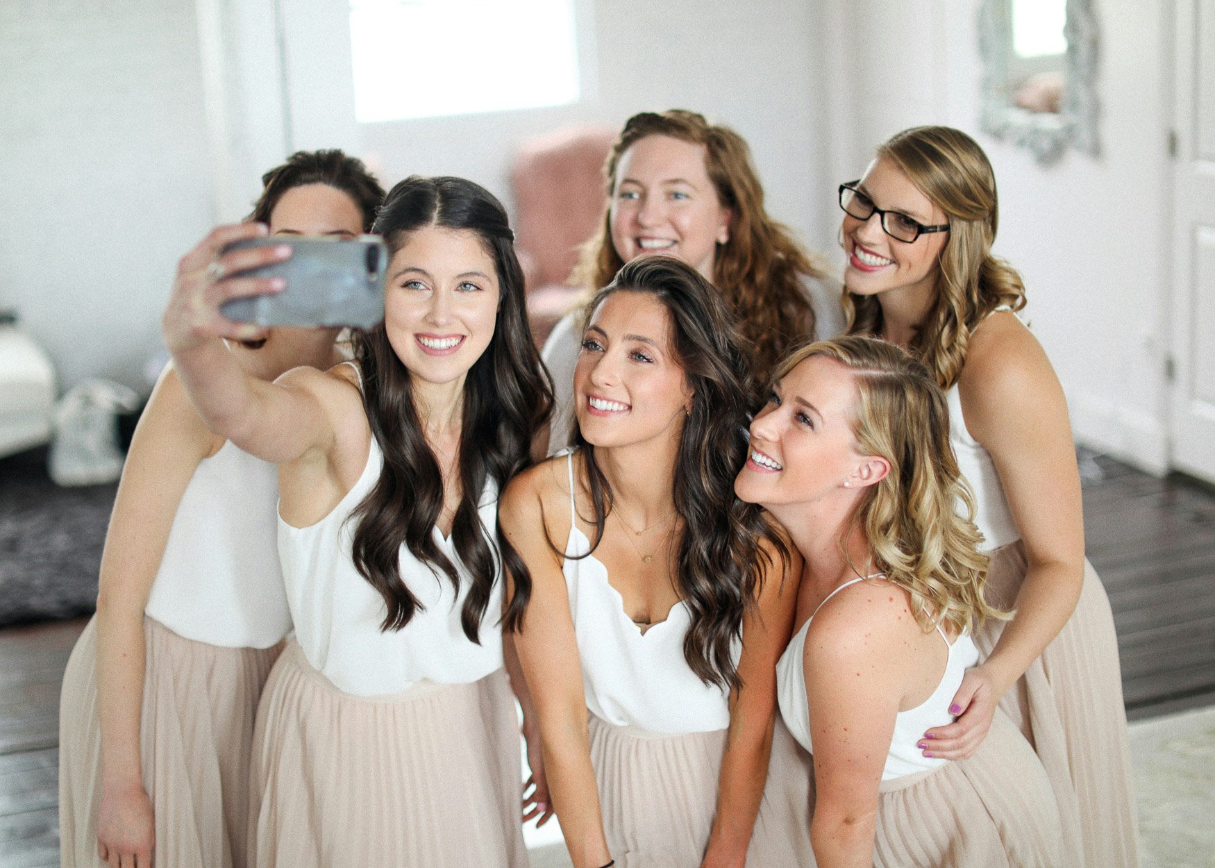 Six bridesmaids in white tops and champagne skirts crowd together for a group selfie in a bright dressing room.