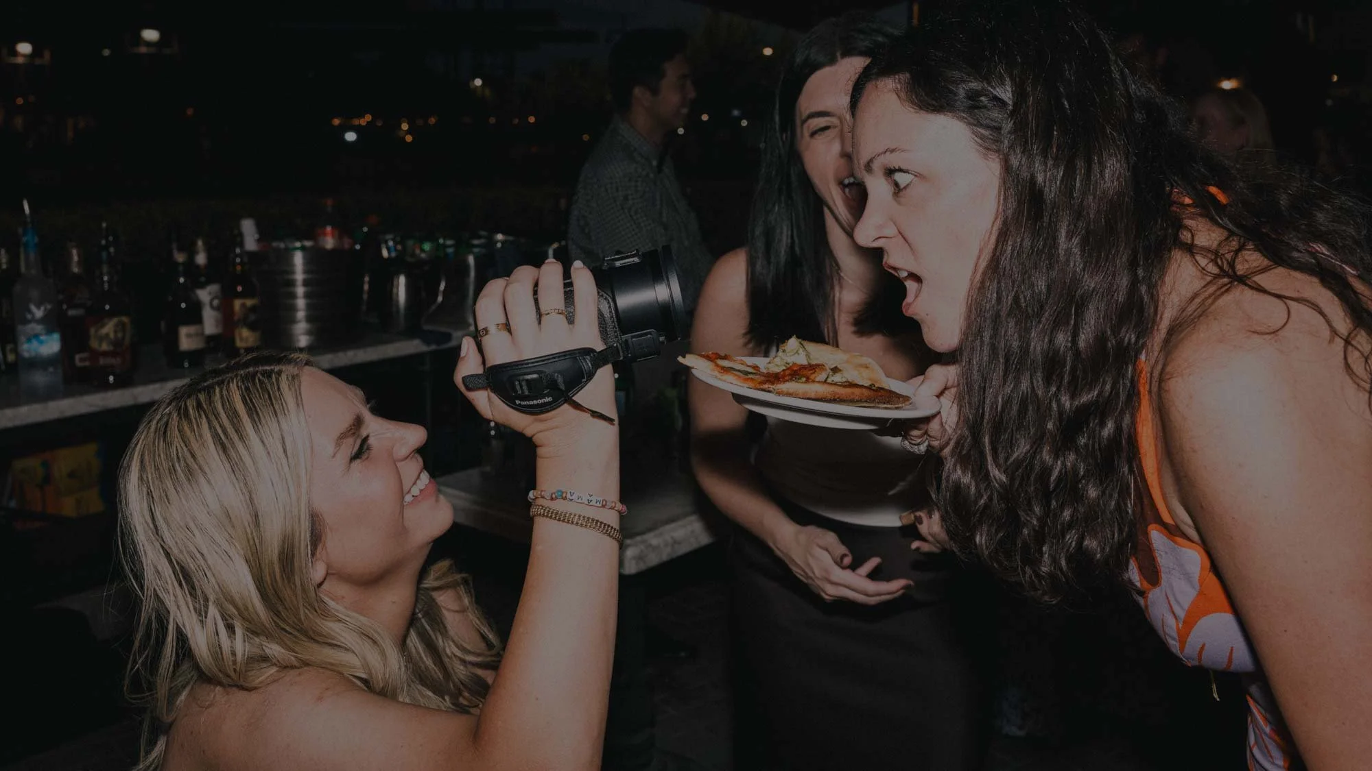A woman films with a camcorder a friend preparing to eat a pizza slice  at a lively nighttime event. She is capturing unscripted moments on video.