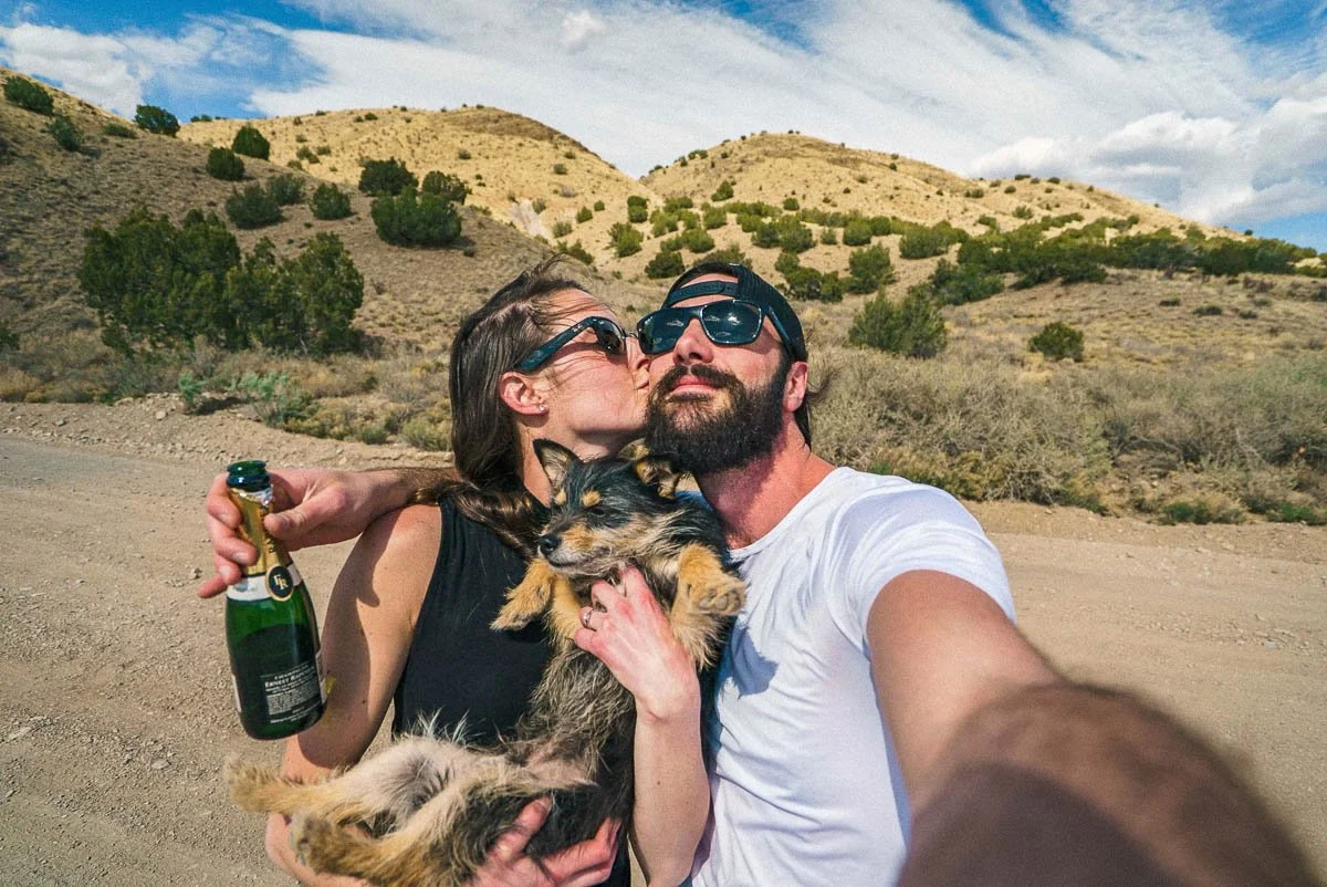 A couple taking a selfie in a desert landscape, with a bottle of Champagne, celebrating an engagement, holding a small dog.