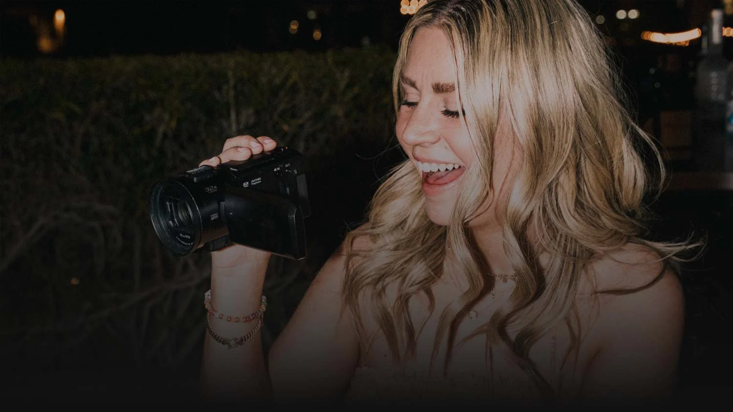 a young woman films on a camcorder at a wedding reception. She is laughing as she records.