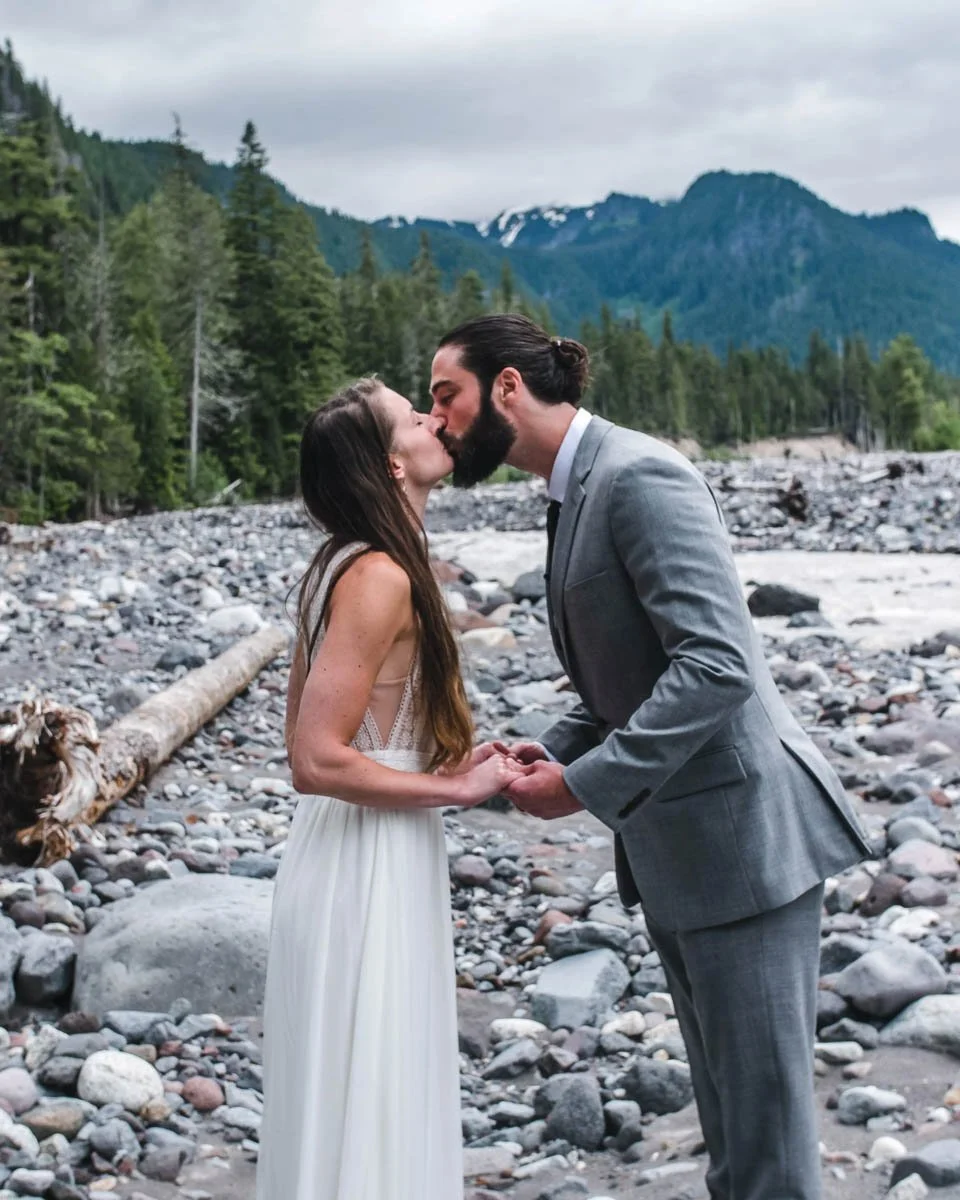 A couple in wedding attire kissing on a rocky riverbank with Mt Rainer and trees in the background.