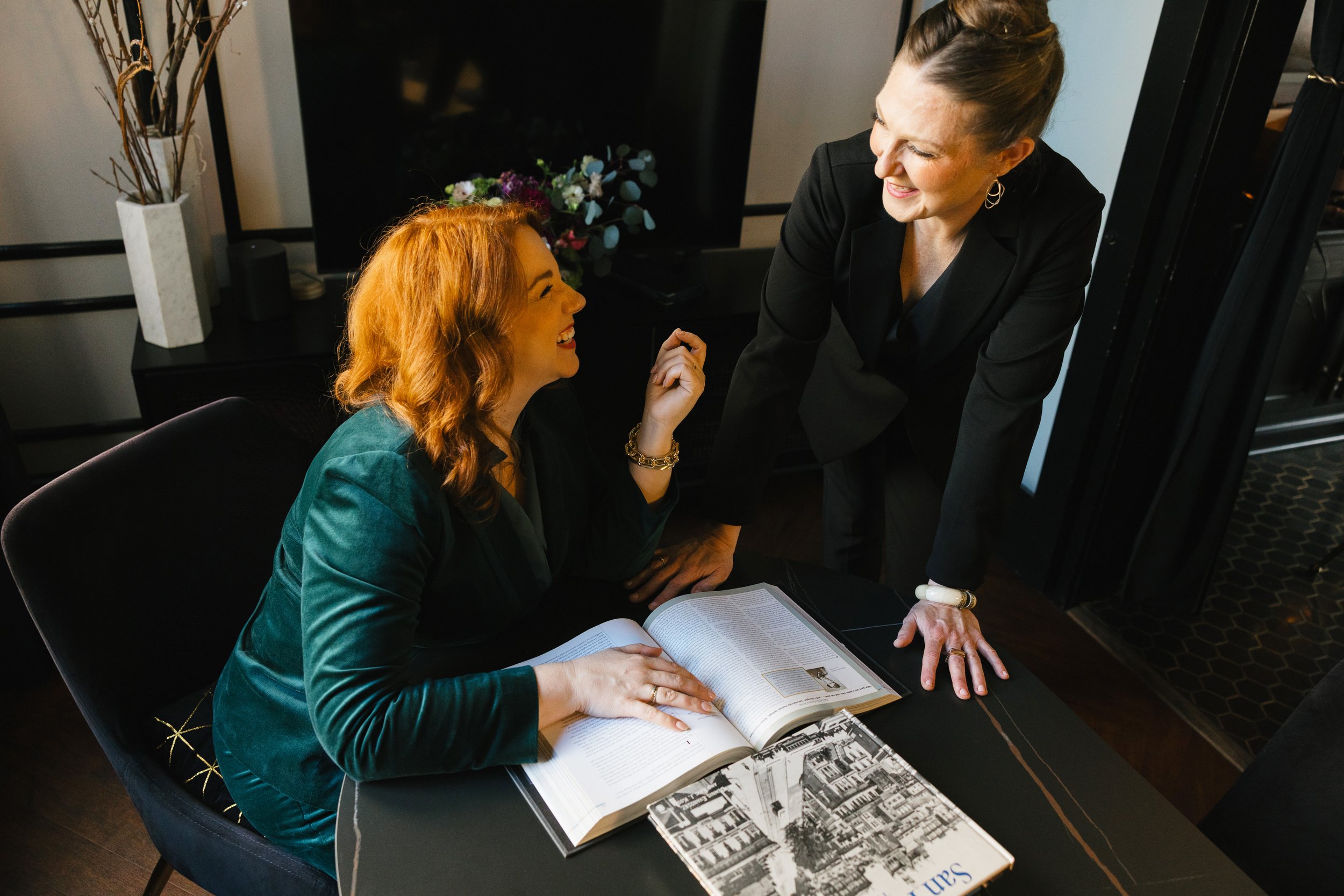 Casey Erin Clark (red hair, seated) and Julie Fogh (dark blonde hair standing), cofounders of Vital Voice Training, engage in a lively discussion in a stylish conference room.