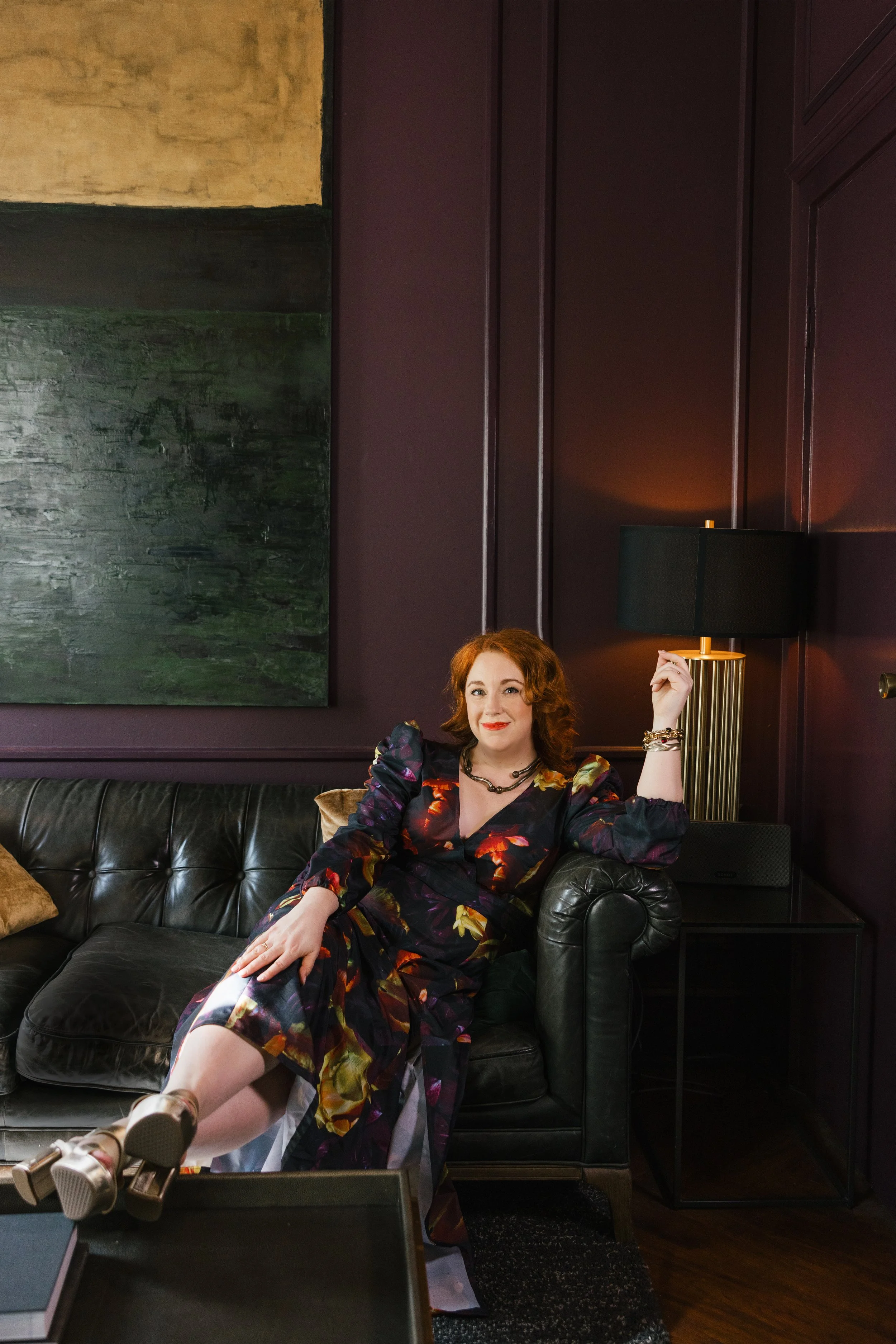 A woman with red hair sitting on a black leather sofa in a dimly lit room with purple walls. She is wearing a colorful dress and jewelry, and is smiling while leaning back with one arm resting on the sofa's armrest and the other raised near a black lampshade on a gold-toned lamp.