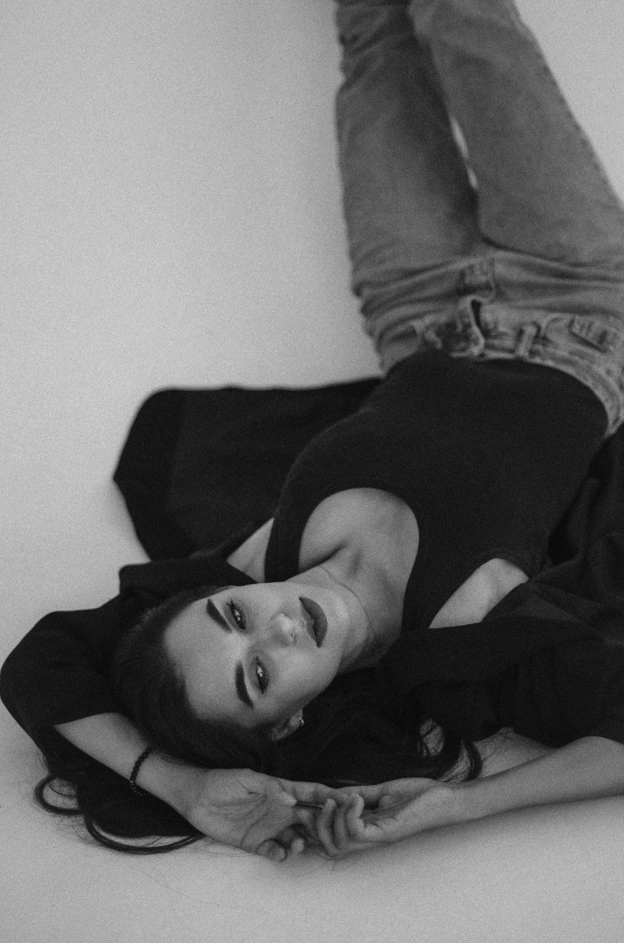 A woman with dark hair lying on her back on a plain surface, wearing a black top and jeans, looking directly at the camera.