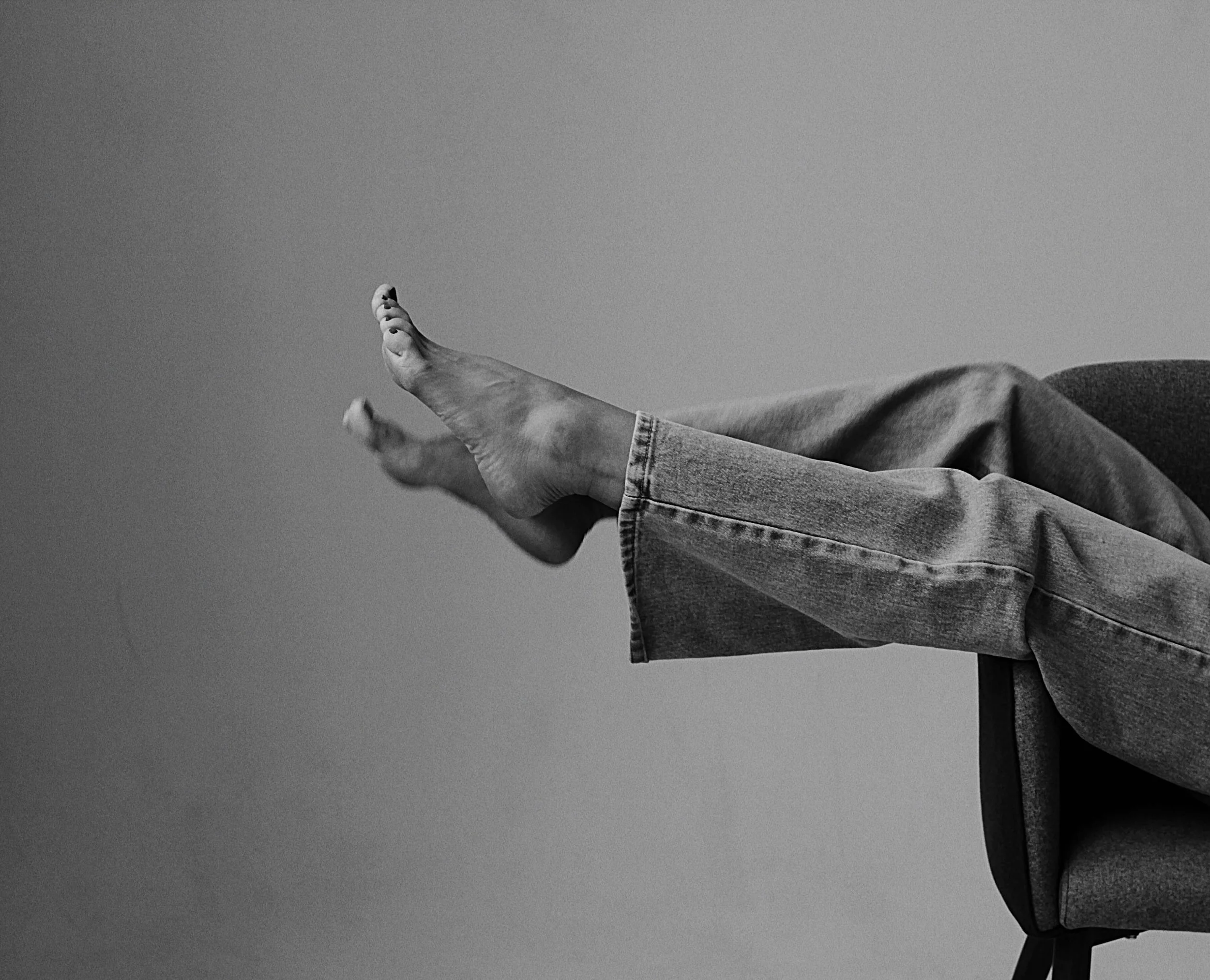 Black and white photo of a person with jeans sitting on a chair, with their legs extended and resting on the armrest.