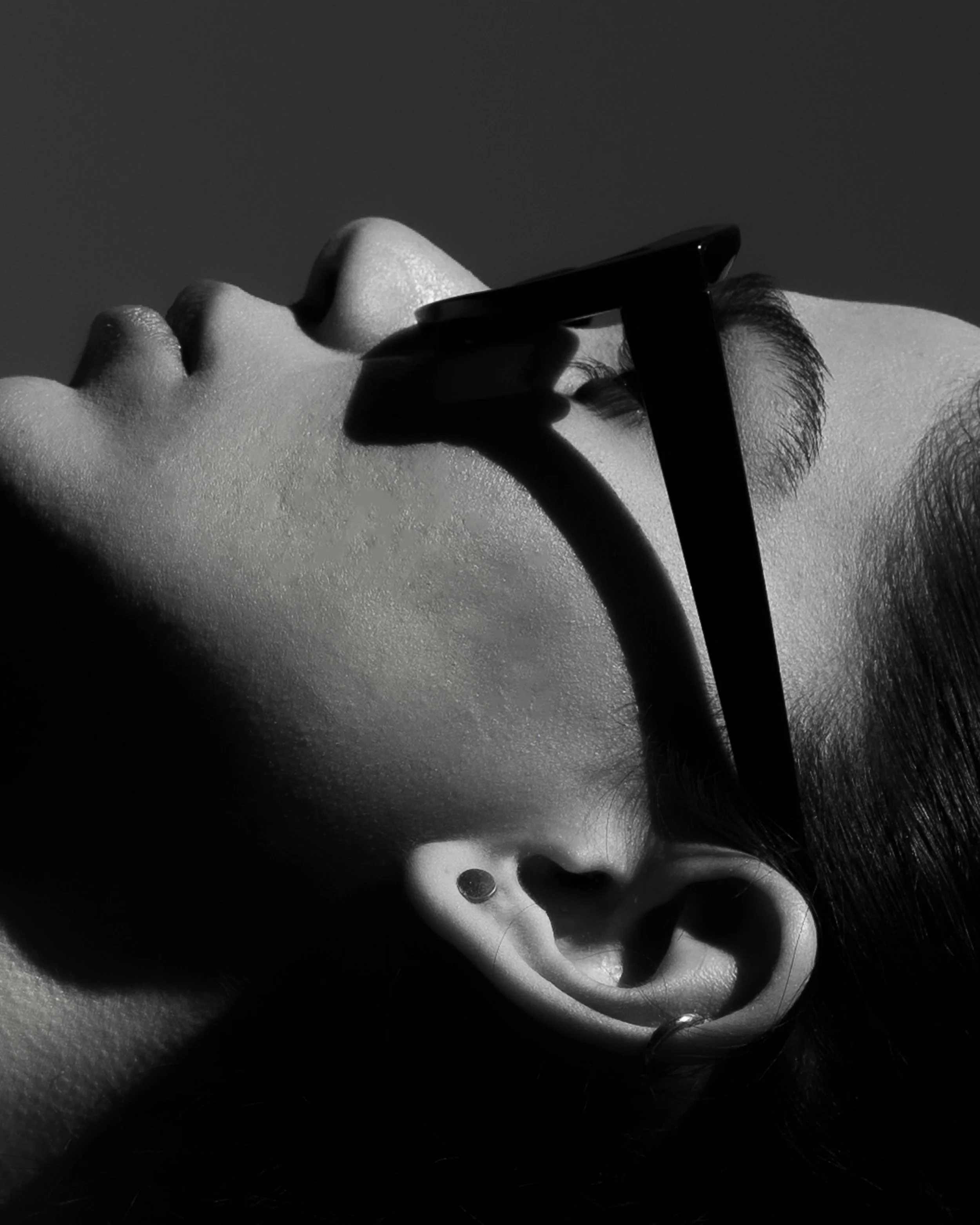 A black and white close-up of a person's face, with glasses resting on the nose, their head tilted back and eye closed, showing an ear with earrings.