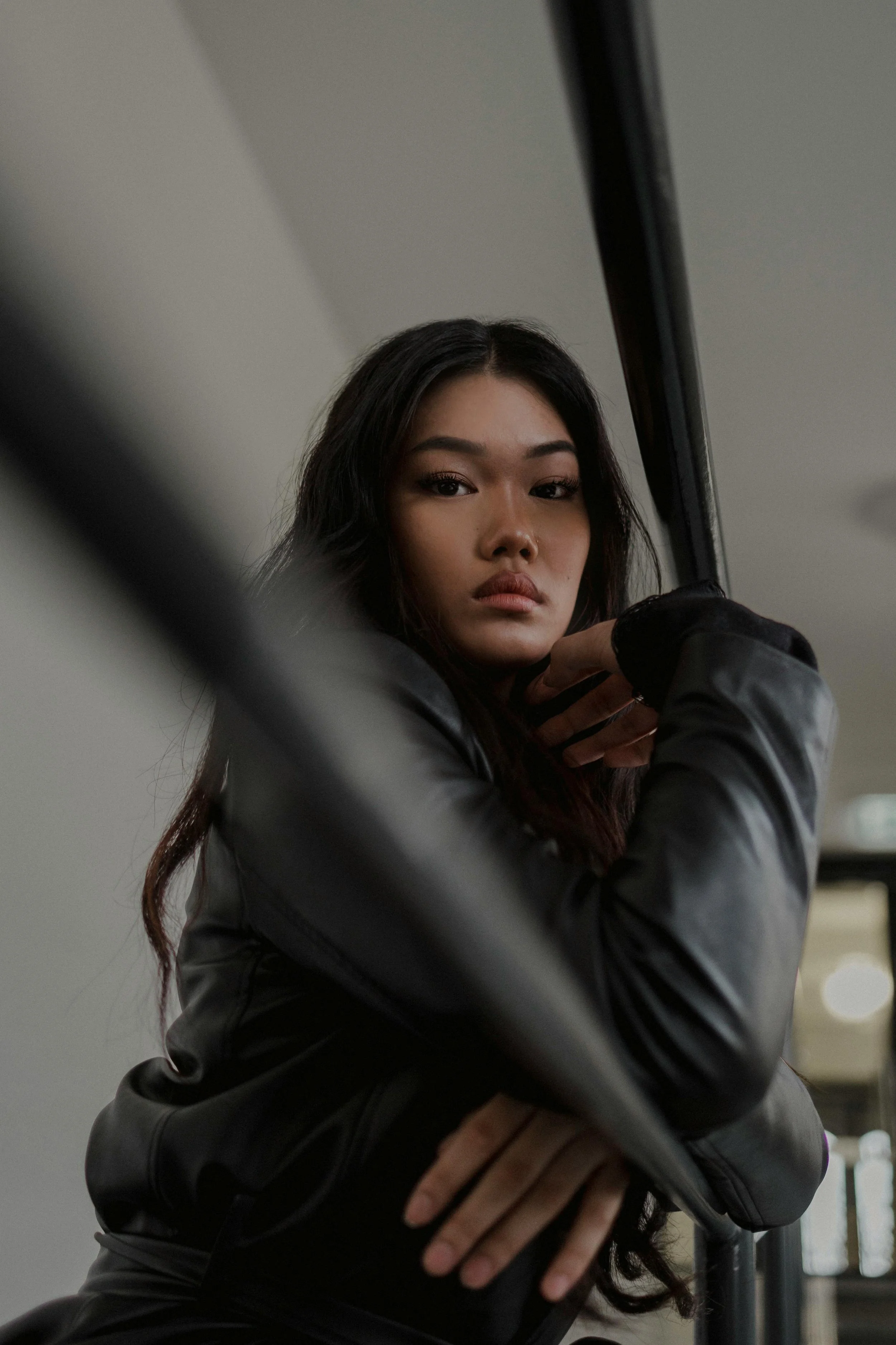 A young woman with long dark hair and tan skin looking at the camera, wearing a black leather jacket, indoors near a mirror or glass panel.