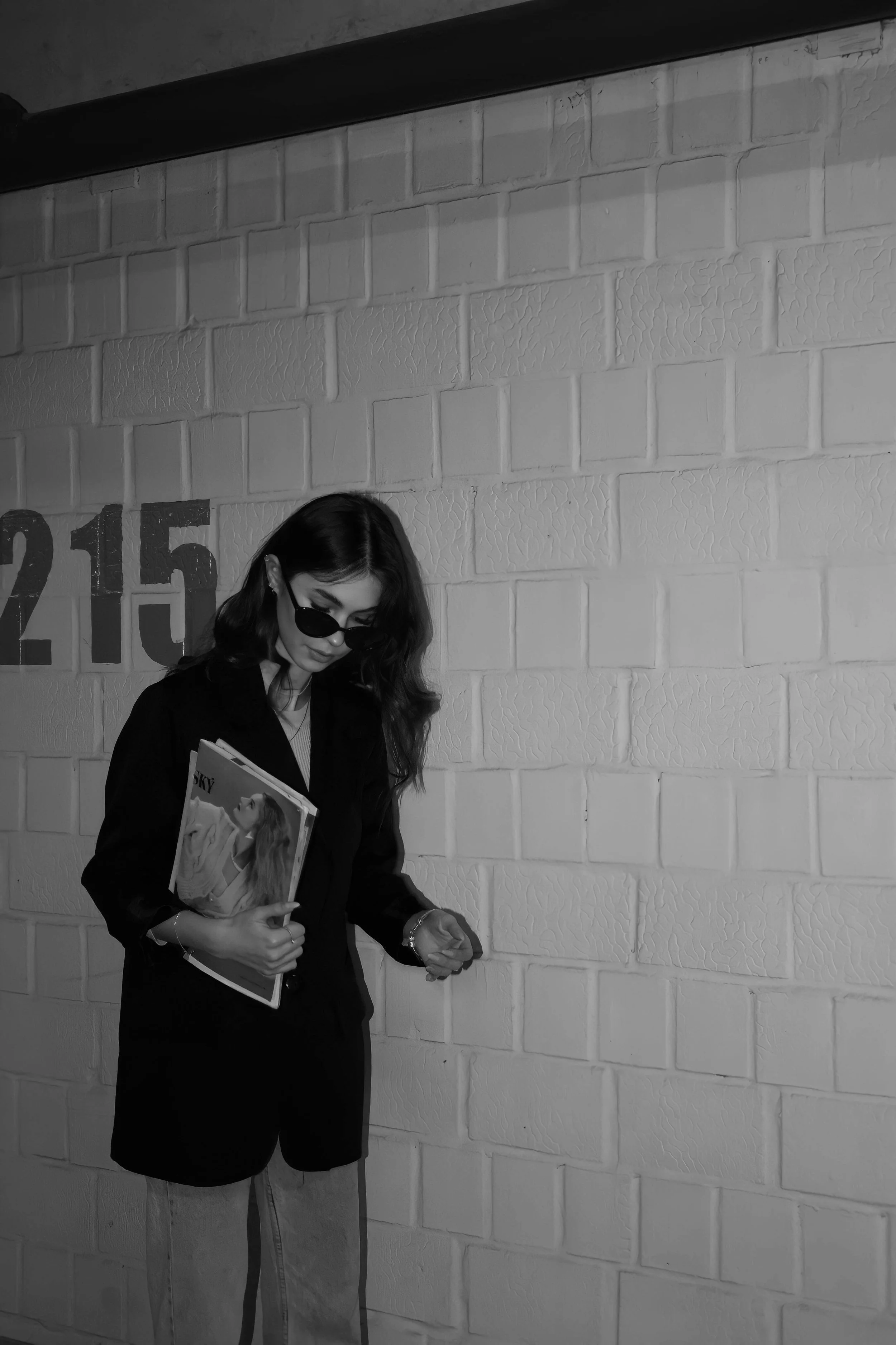 A woman with long dark hair and sunglasses, holding magazines, standing against a white brick wall with the number 215 partially visible. She is wearing a black blazer and light-colored pants.