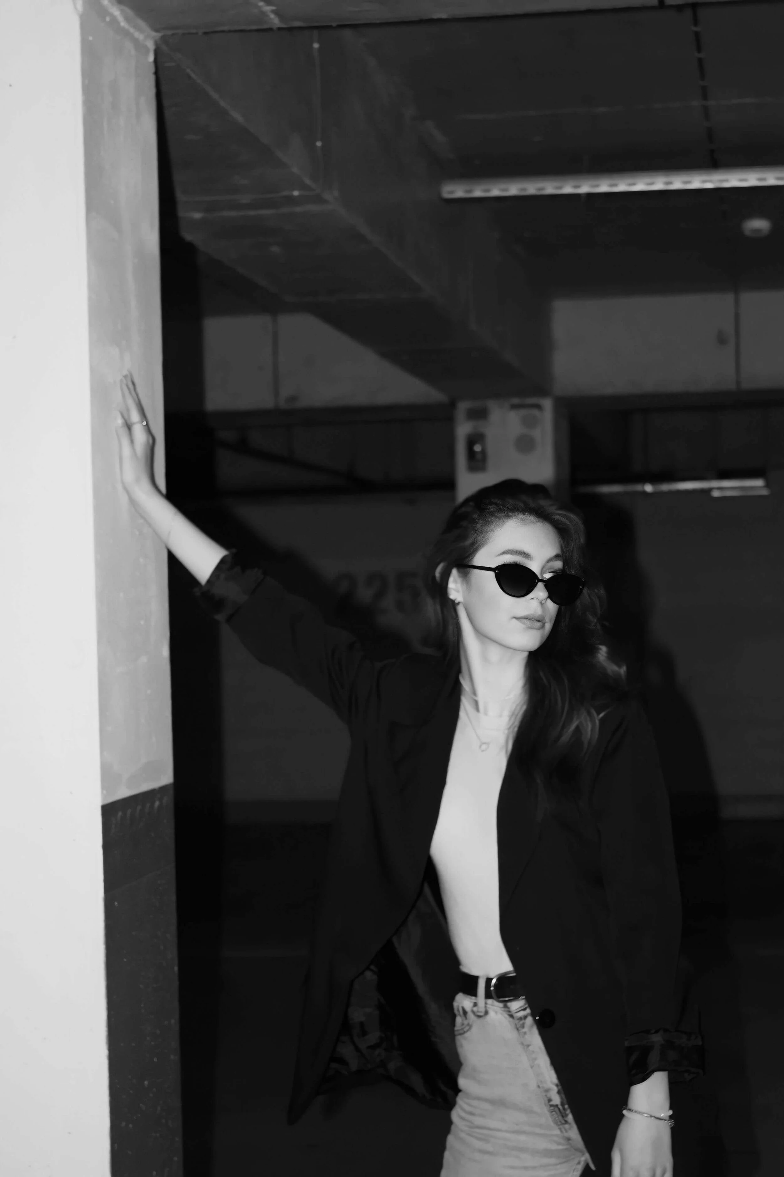A woman with long hair wearing sunglasses, a black blazer, light-colored top, and jeans, standing in a dimly lit indoor parking garage with her arm touching a column.