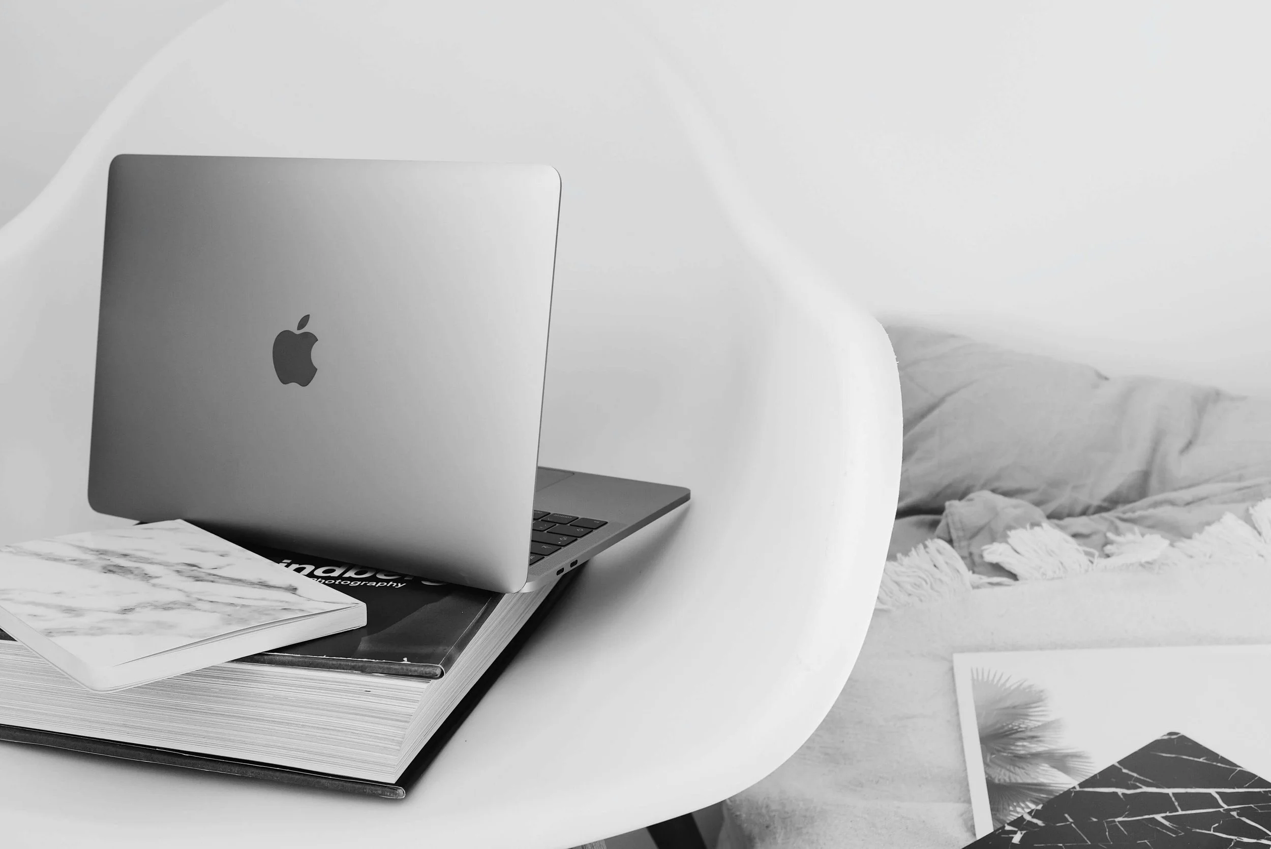 Black and white photo of a closed MacBook on top of a stack of magazines and books, next to a marble-patterned notebook, with a bed and pillow in the background.