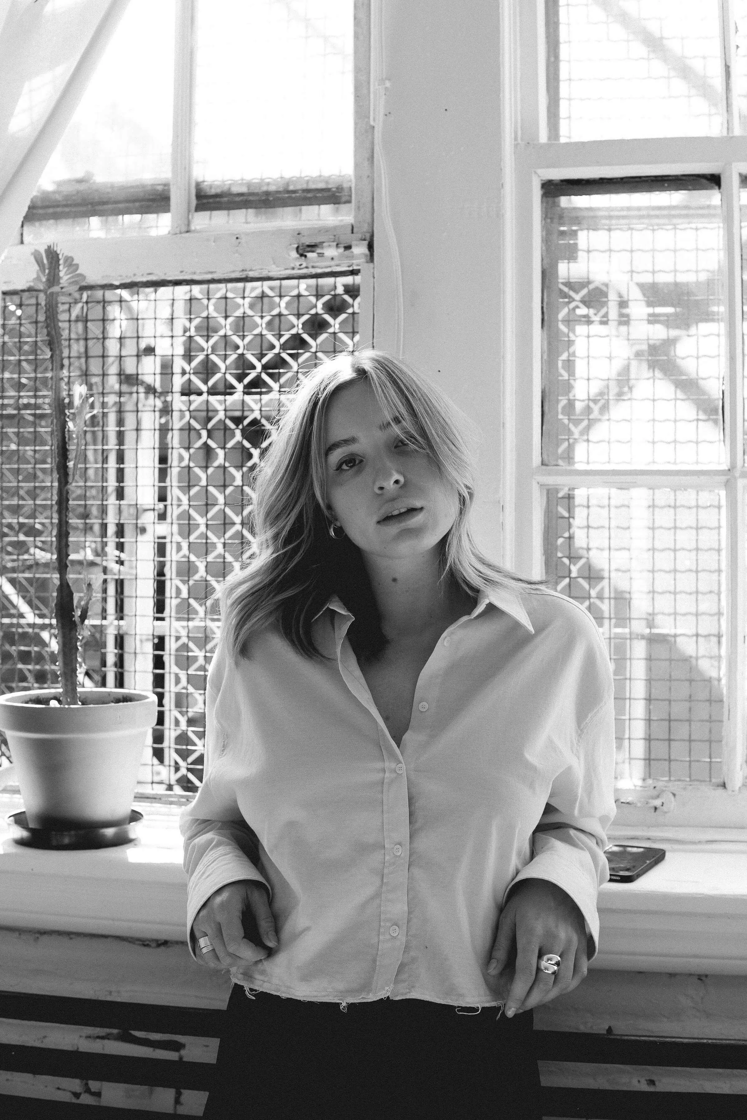 A young woman with shoulder-length hair, wearing a white button-up shirt, stands indoors in front of a window with metal grid and wire mesh security, with sunlight streaming in. A potted cactus is on the windowsill, and a smartphone rests nearby.