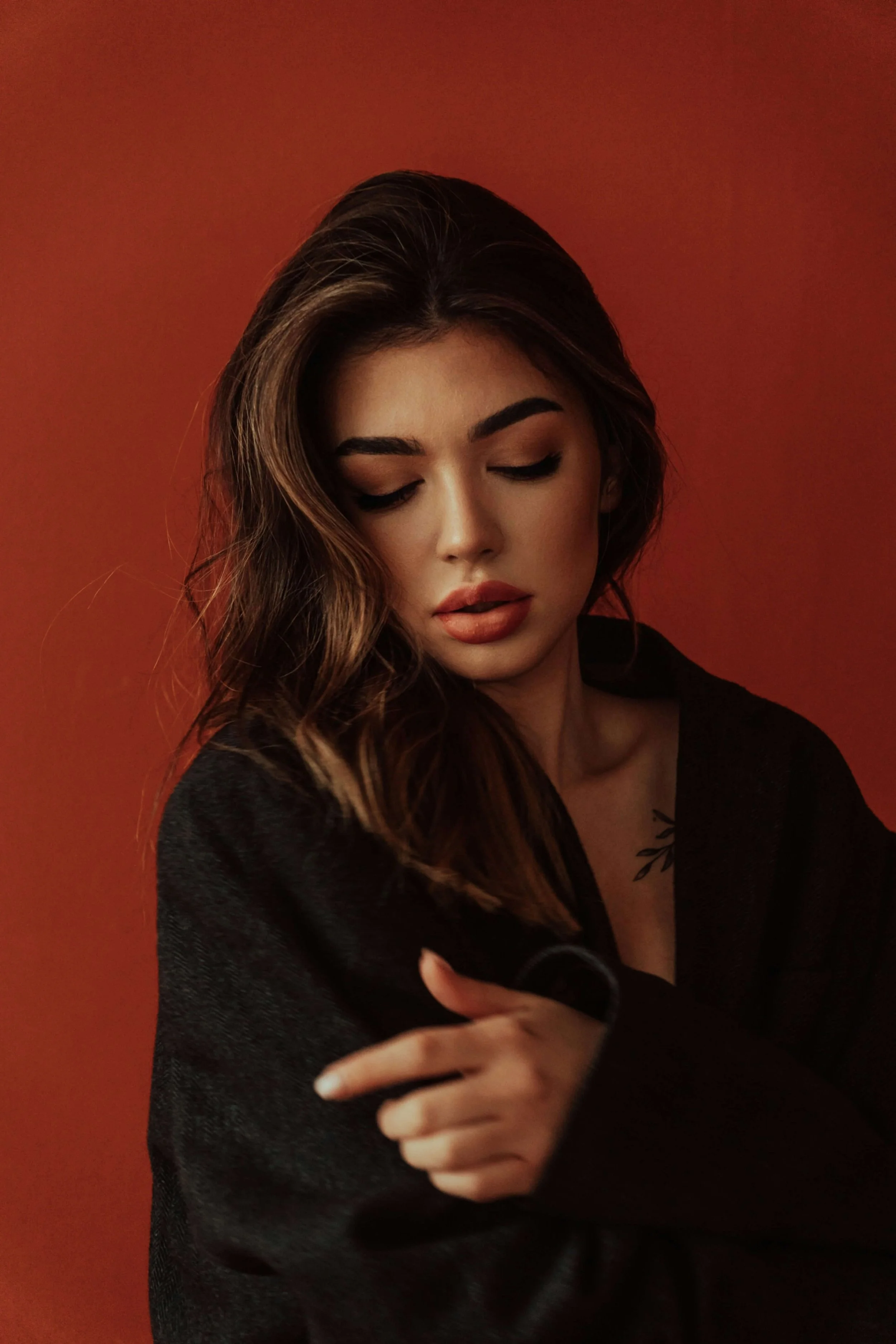 Young woman with wavy brown hair, closed eyes, and natural makeup, wearing a black jacket against a red background.