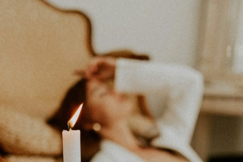 A woman lying on a bed taking a selfie with her phone while a lit white candle sits in the foreground.