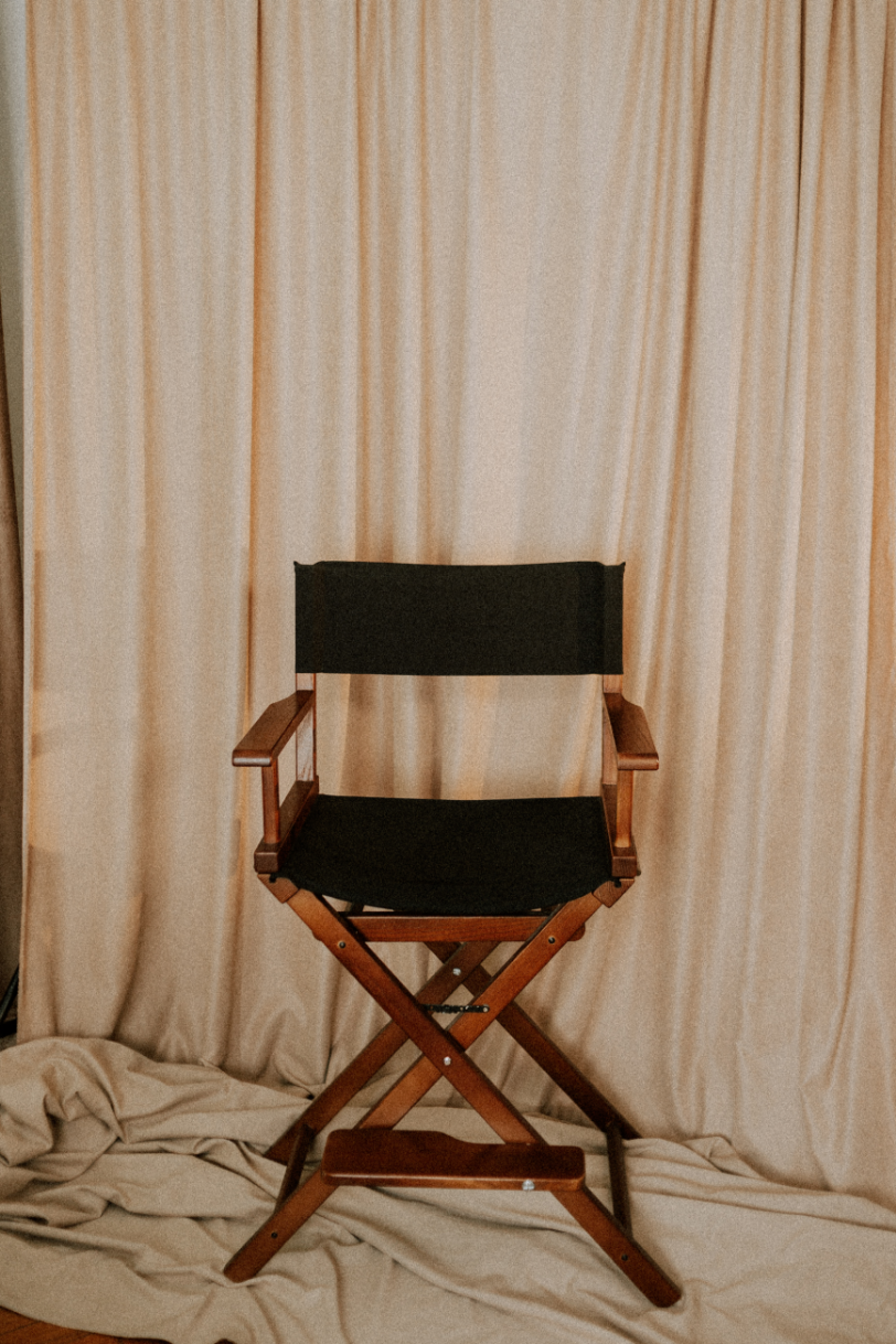 A director's chair with black fabric and wooden armrests, placed on a beige fabric backdrop.