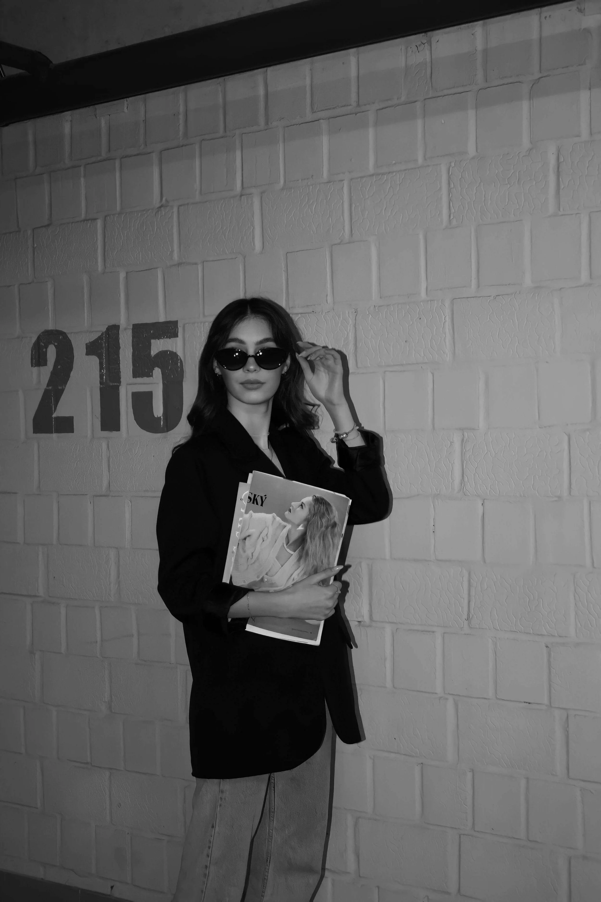 A woman with shoulder-length hair wearing sunglasses, a black blazer, and light-colored jeans standing against a brick wall with the number 215 painted on it. She is holding magazines or photos, and adjusting her sunglasses.