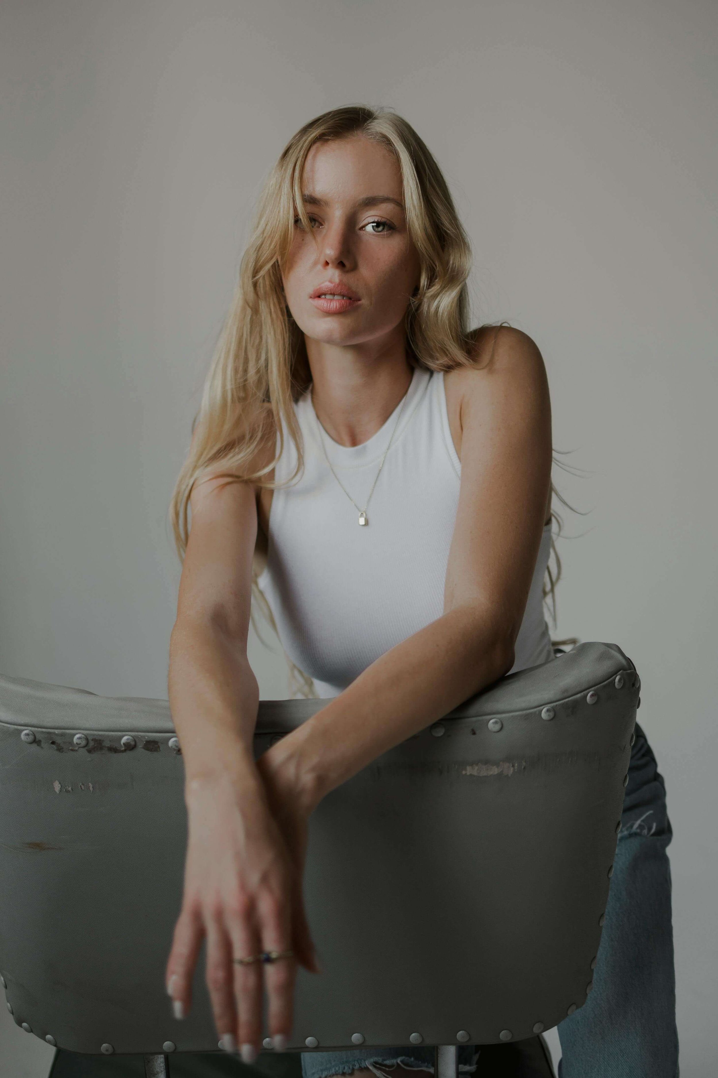 A young woman with long wavy blonde hair, wearing a white sleeveless top and jeans, leaning forward on a gray chair with rivet details, against a plain light background.