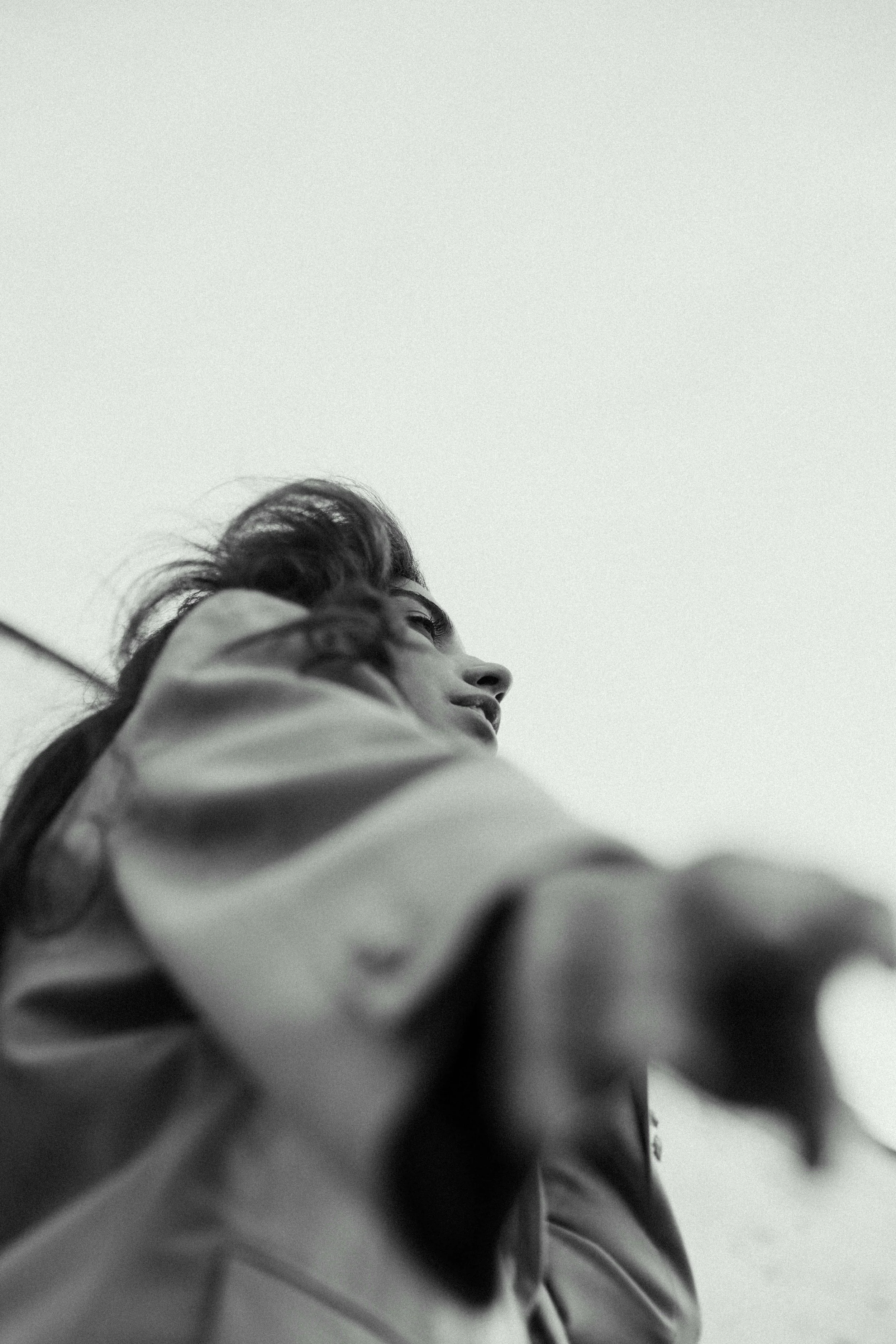 A black and white photo of a woman looking up, with her face turned sideways and her hair blowing in the wind, wearing a jacket.
