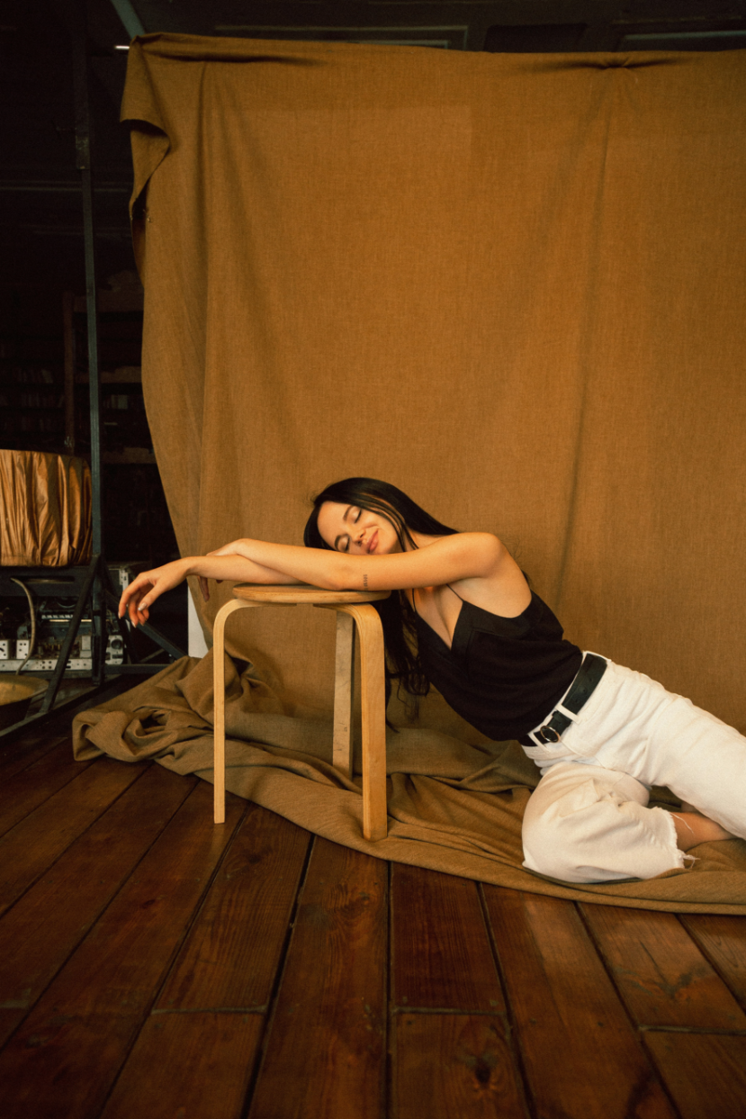 A woman with long dark hair and tattoos, wearing a black tank top and white pants, is sitting on a wooden floor, leaning against a small wooden table. She is smiling with her eyes closed, resting her head and arms on the table in a relaxed pose. Behind her is a large beige fabric backdrop, and the setting appears to be indoors with studio lighting.