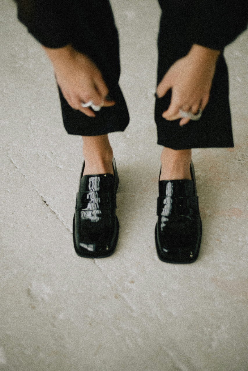 Person wearing black shoes with a shiny finish, black pants, and standing on a beige tiled floor.