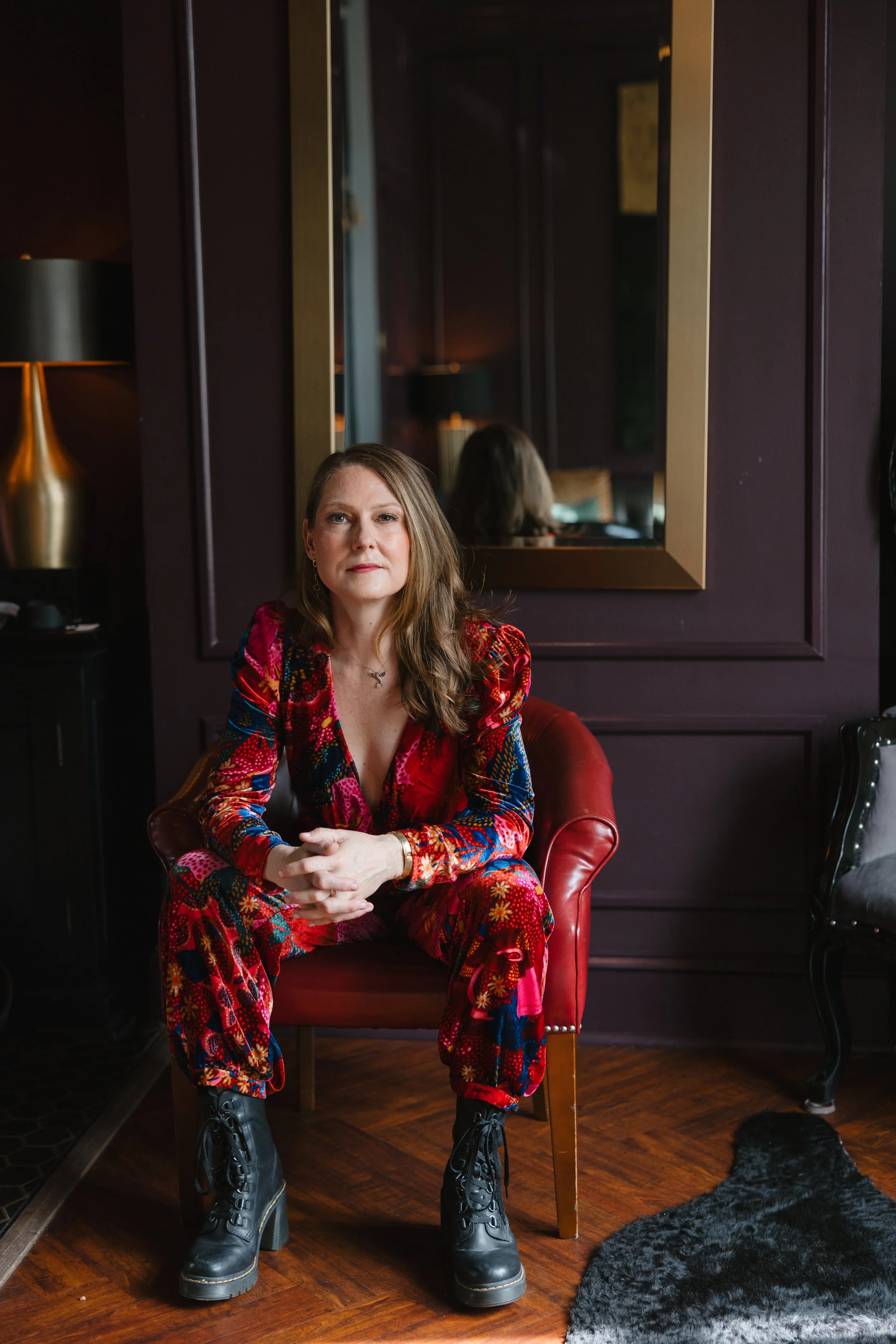 A woman sitting on a red leather armchair in a dimly lit room, wearing a colorful, patterned outfit and black lace-up boots, with a large mirror reflecting her back and the dark purple walls behind her.