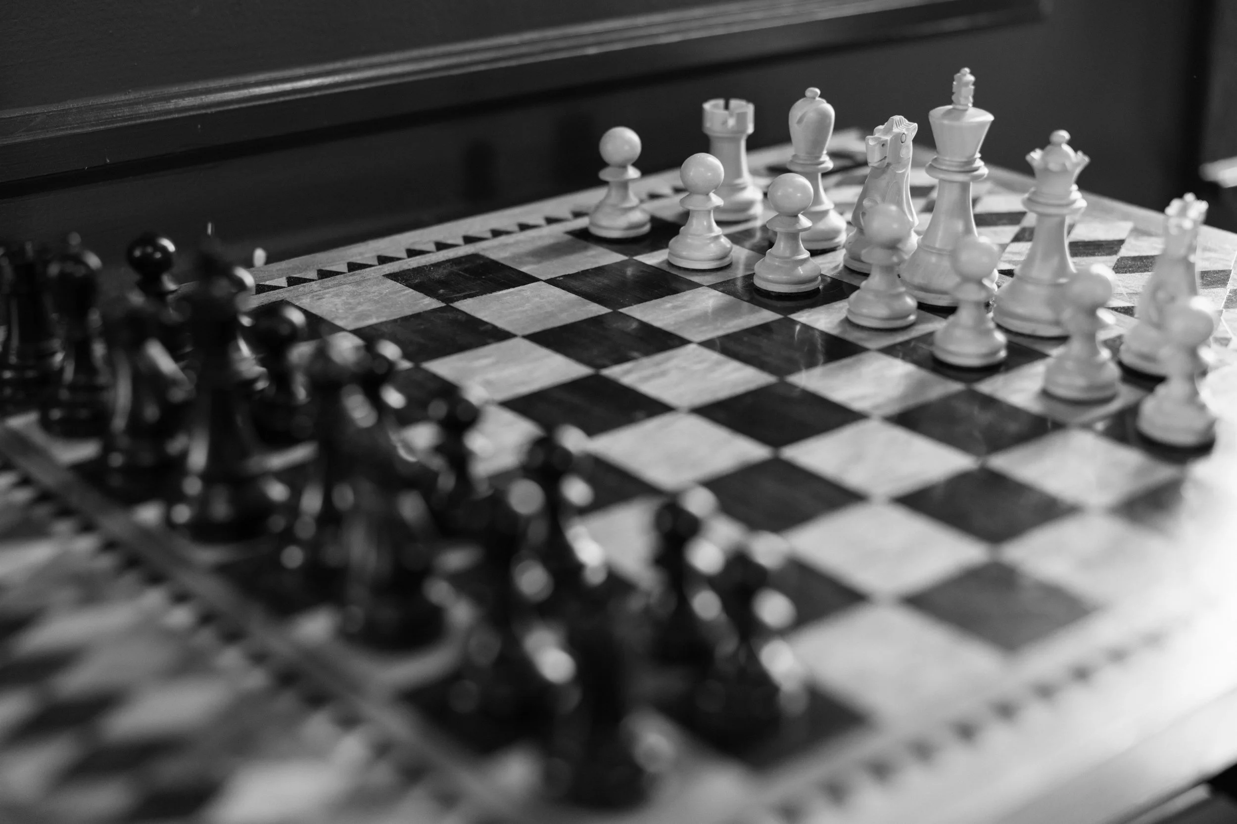 A chessboard with chess pieces arranged for a game, in black and white, with the focus on the white pieces and the black pieces blurred in the foreground.