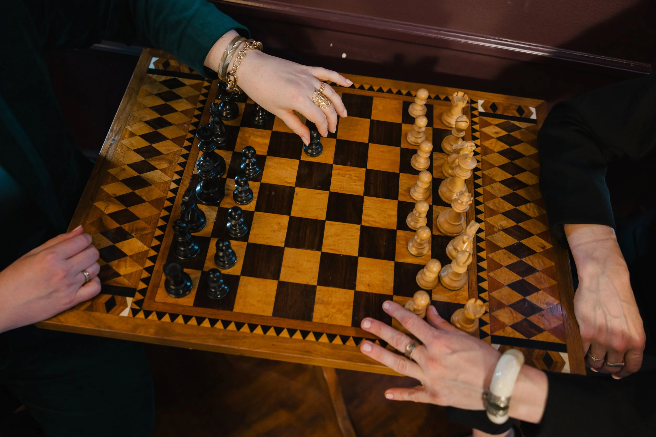Two women playing chess at a wooden chessboard; one woman's hand is moving a black piece, while the other's hand rests on the edge of the board.