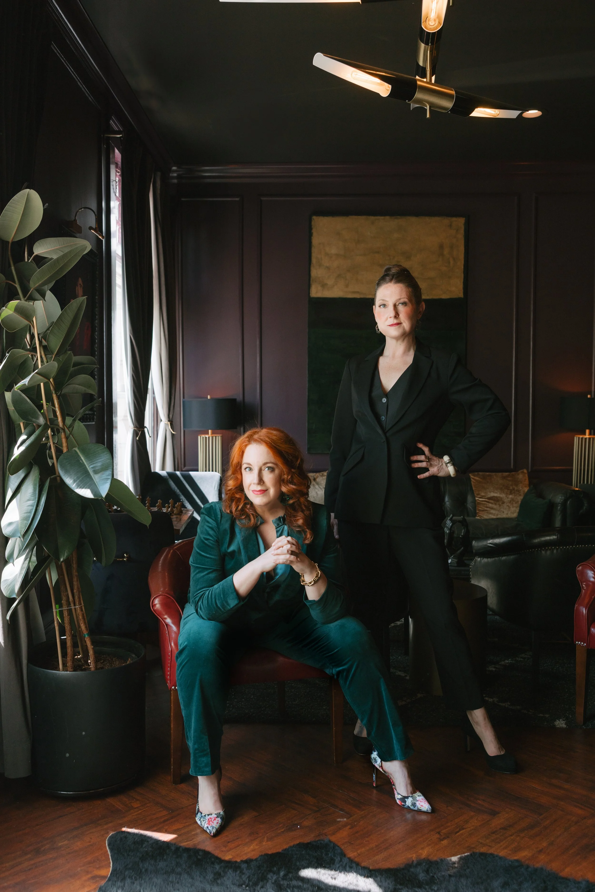 Casey Erin Clark (seated in a dark green velvet suit) and Julie Fogh (standing in a sharp black suit), cofounders of Vital Voice Training, in a dark, elegant room — they look directly at the camera with small smiles as if to say “let’s go!”