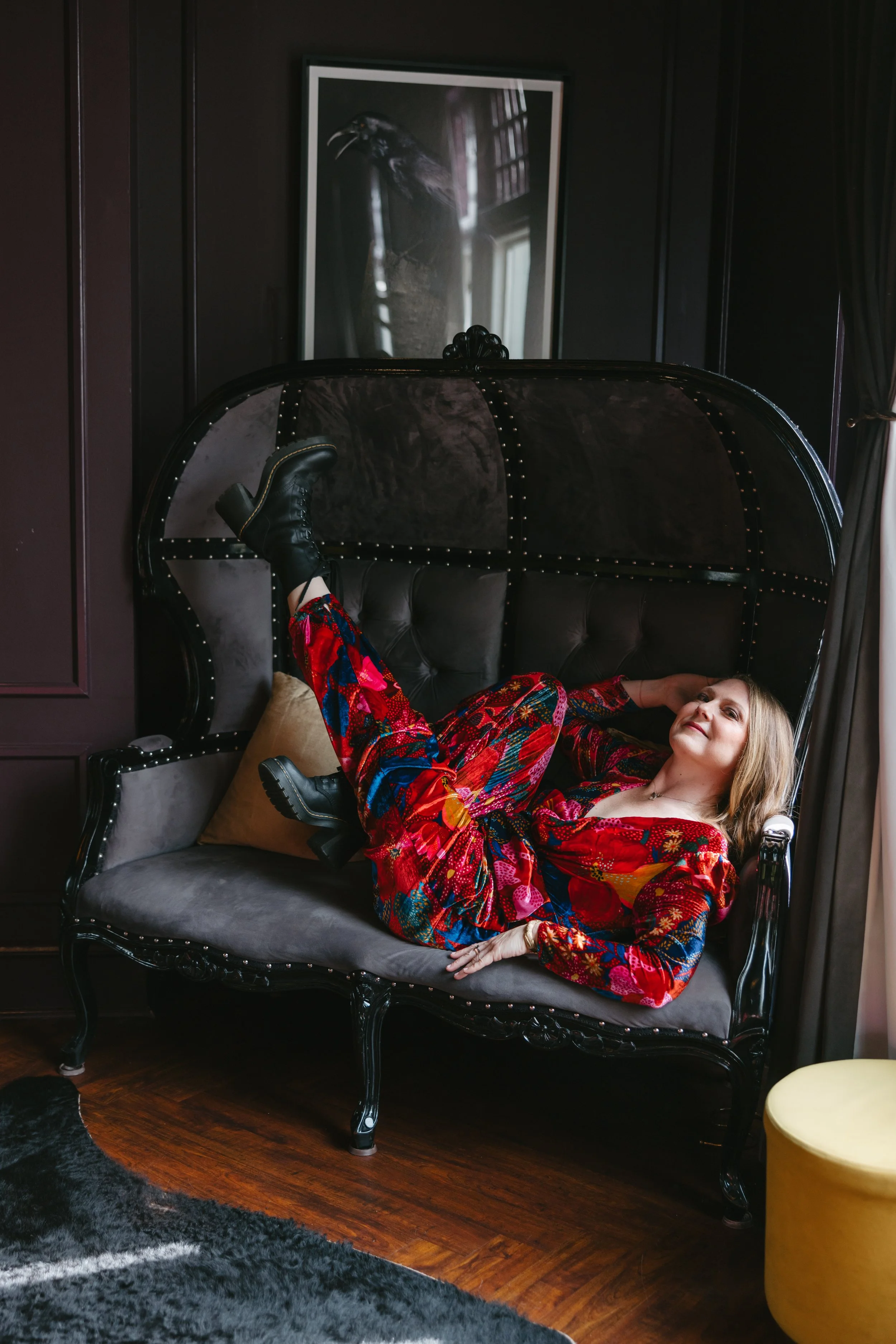 A woman lying on a black vintage sofa with a tufted backrest and curved armrests, wearing colorful patterned clothing and black boots, in a dark room with a framed black and white raven photograph on the wall behind her.