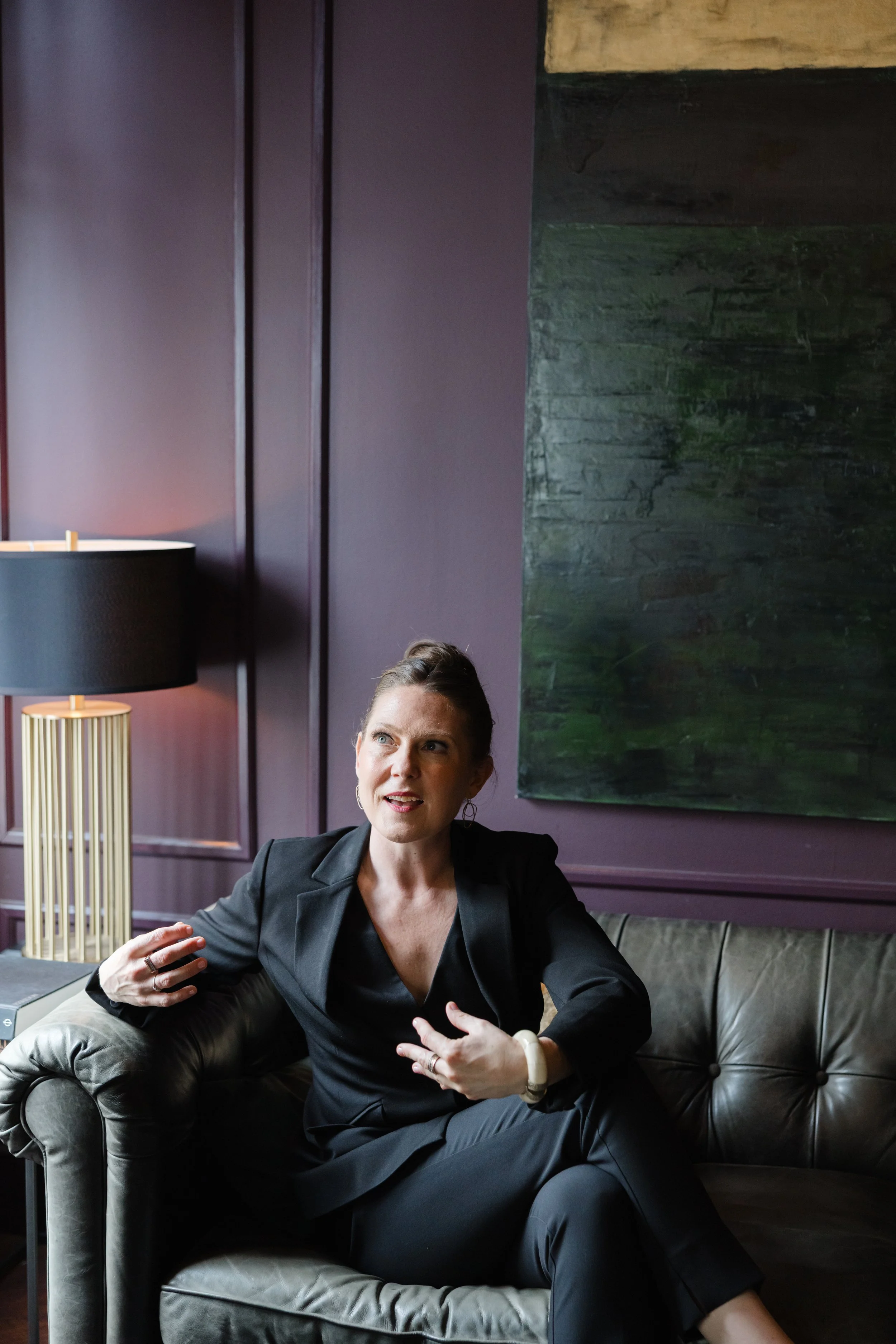 A woman sitting on a dark leather sofa in a room with purple walls and a large dark painting. She is wearing a black suit and has short brown hair. She appears to be engaged in a conversation or explaining something, with one arm resting on the sofa armrest and the other gesturing.