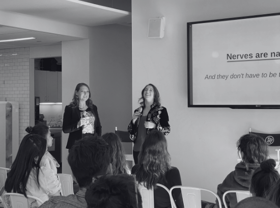 Two women give a presentation to an audience in a room with a large screen displaying partially visible presentation slide.
