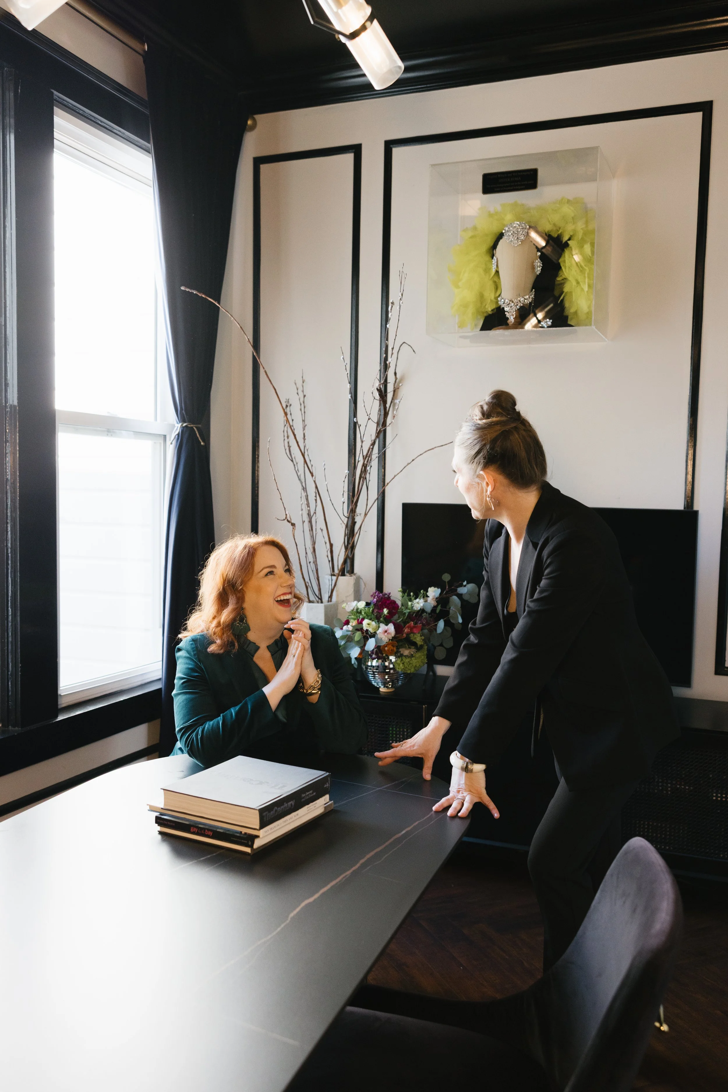 Two women having a joyful conversation in a modern office, one sitting at a desk with books, the other standing and leaning on the table, with a flower arrangement and artistic decor in the background.