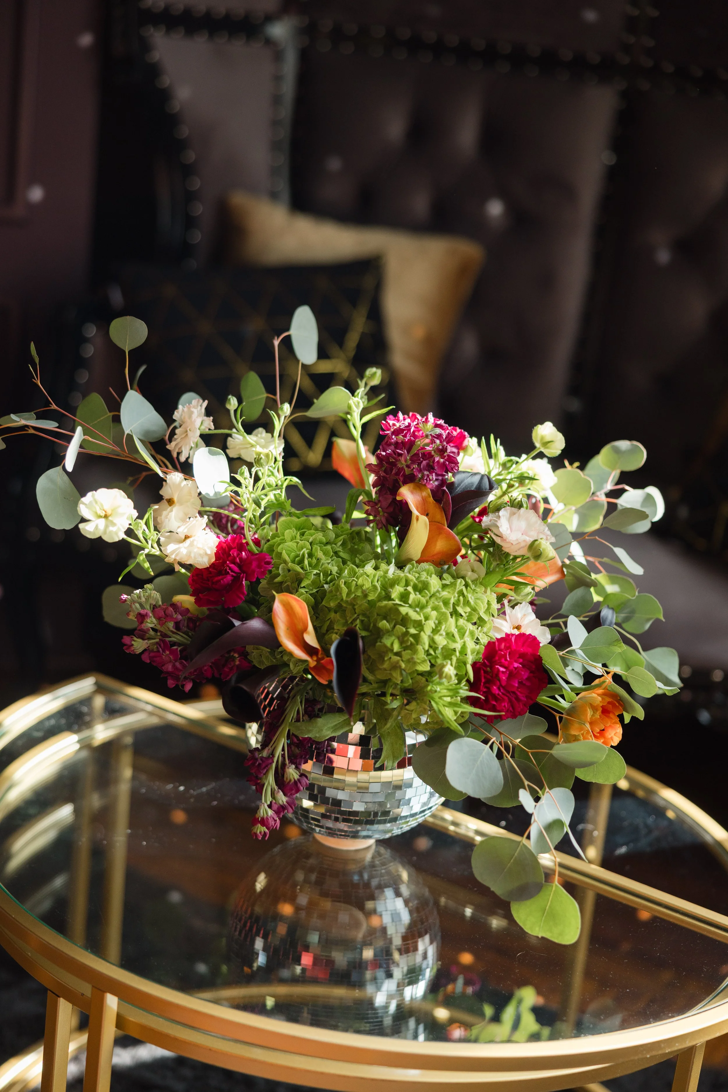 A vibrant flower arrangement in a mosaic vase on a gold-framed glass table inside a dimly-lit room.