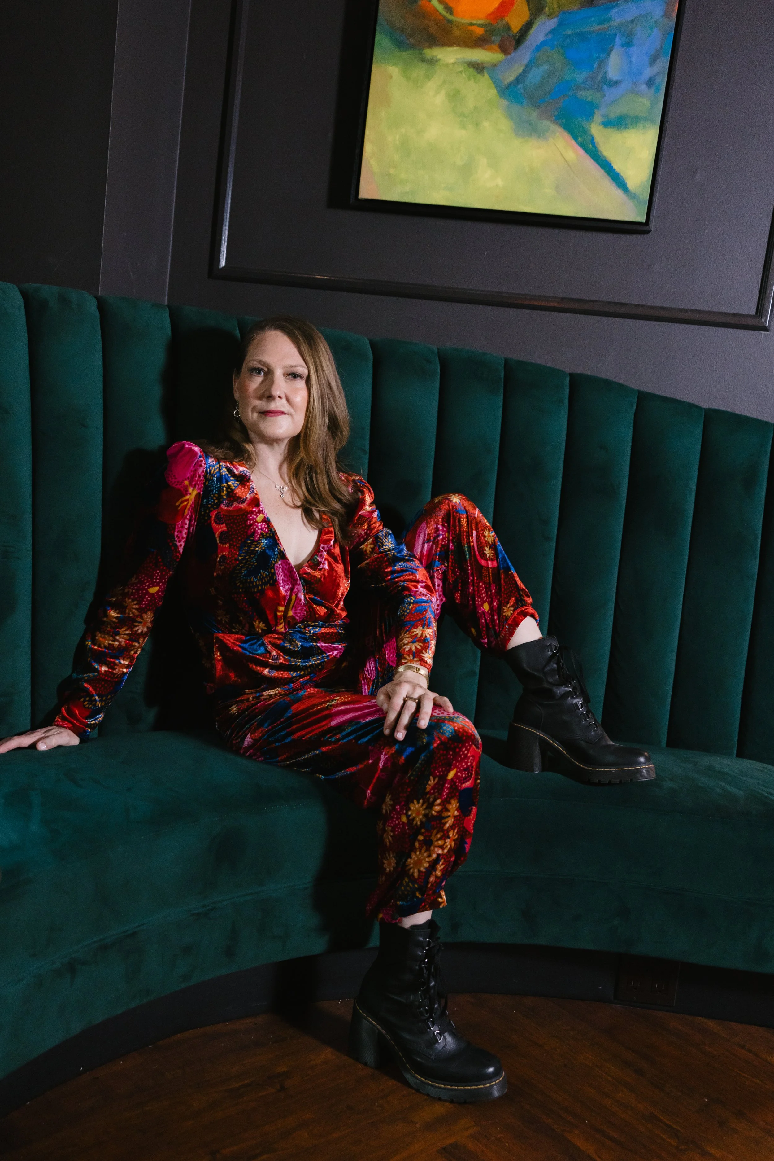 Julie Fogh, cofounder of Vital Voice Training, in a colorful floral jumpsuit and black lace-up boots sitting on a green velvet curved booth. She looks directly at the camera. Intelligence shines from her eyes.