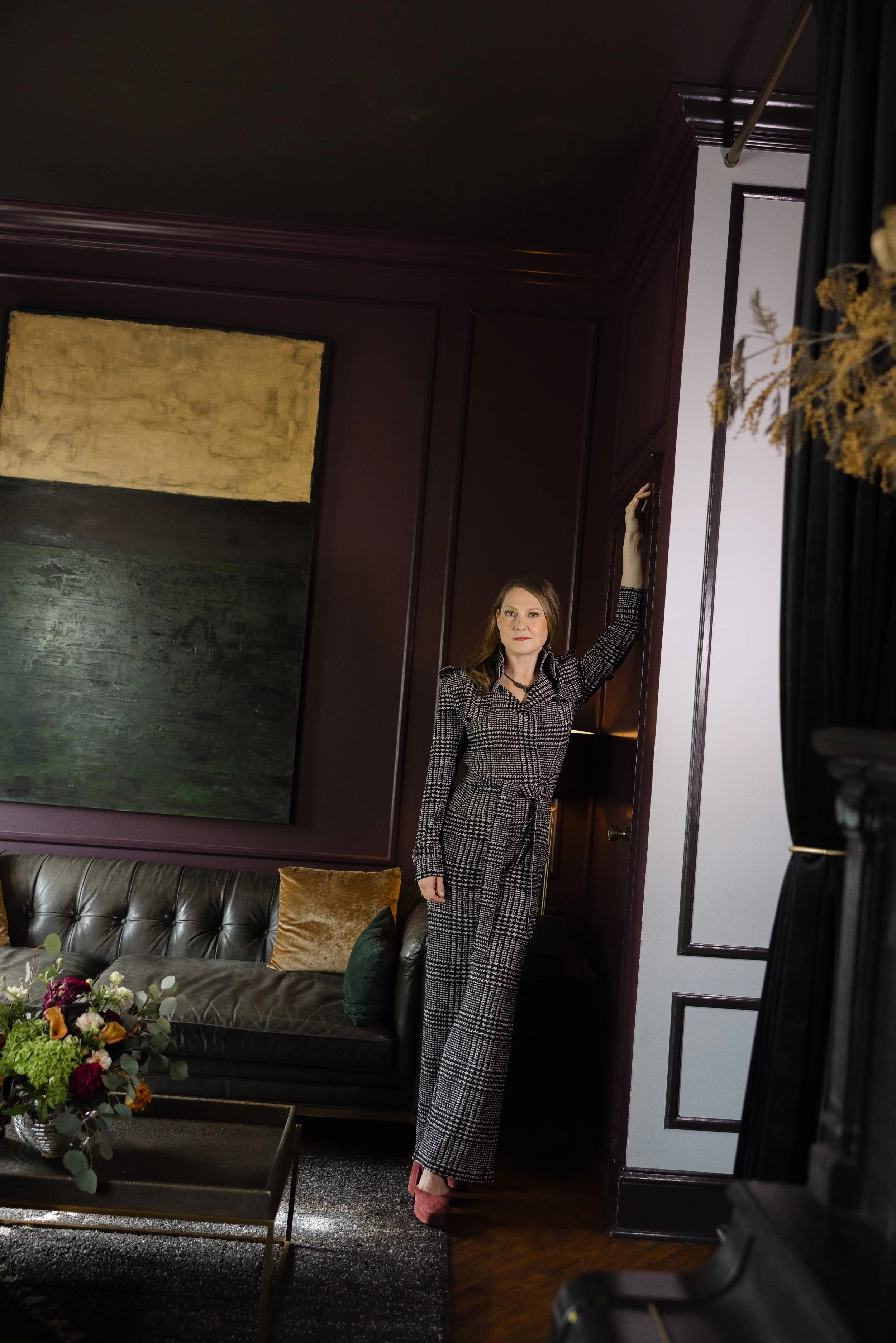 Woman in plaid jumpsuit standing in a living room with dark furniture and artwork, holding a curtain rod.