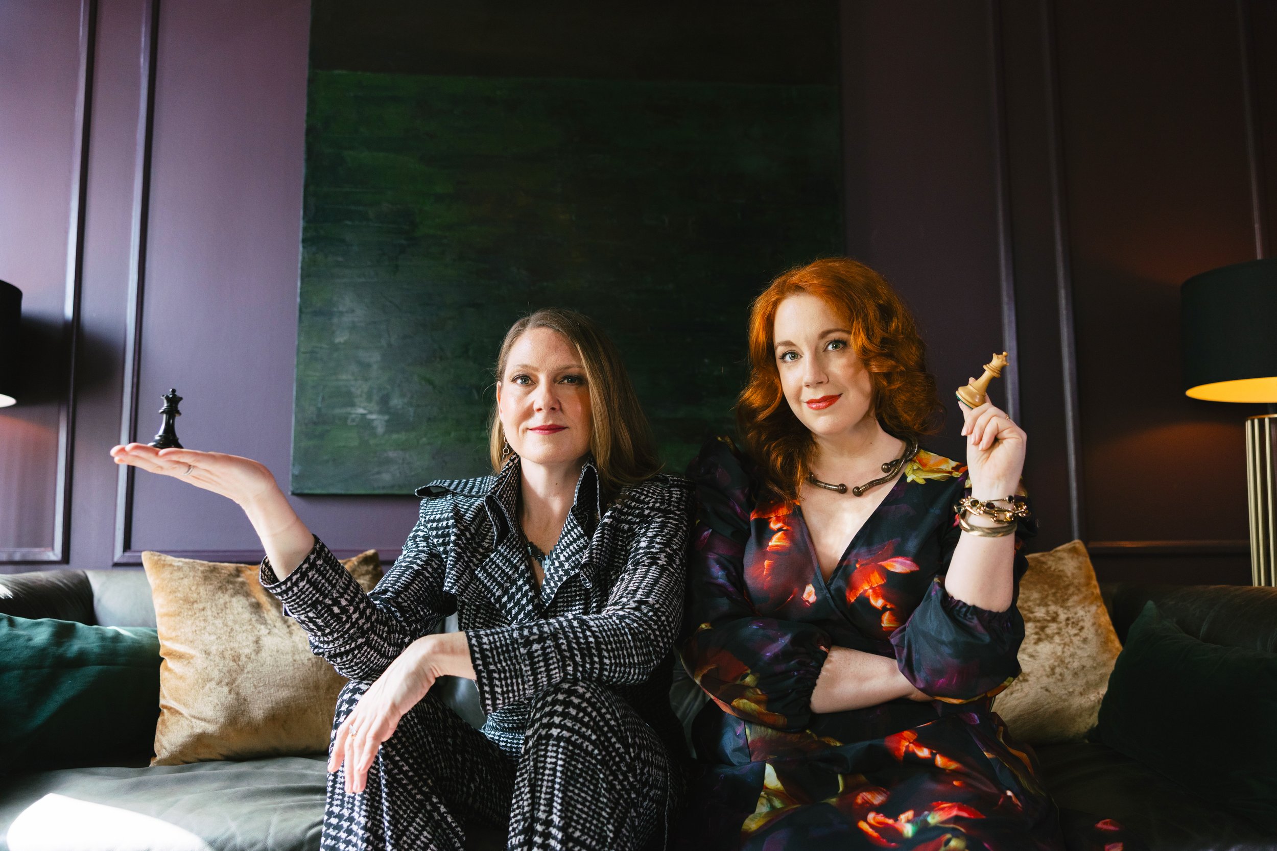 Casey Erin Clark and Julie Fogh, cofounders of Vital Voice Training, sit on a dark-colored sofa in a stylish room, each holding queen chess pieces and smiling slightly at the fewer. They look powerful and a bit mischievous.