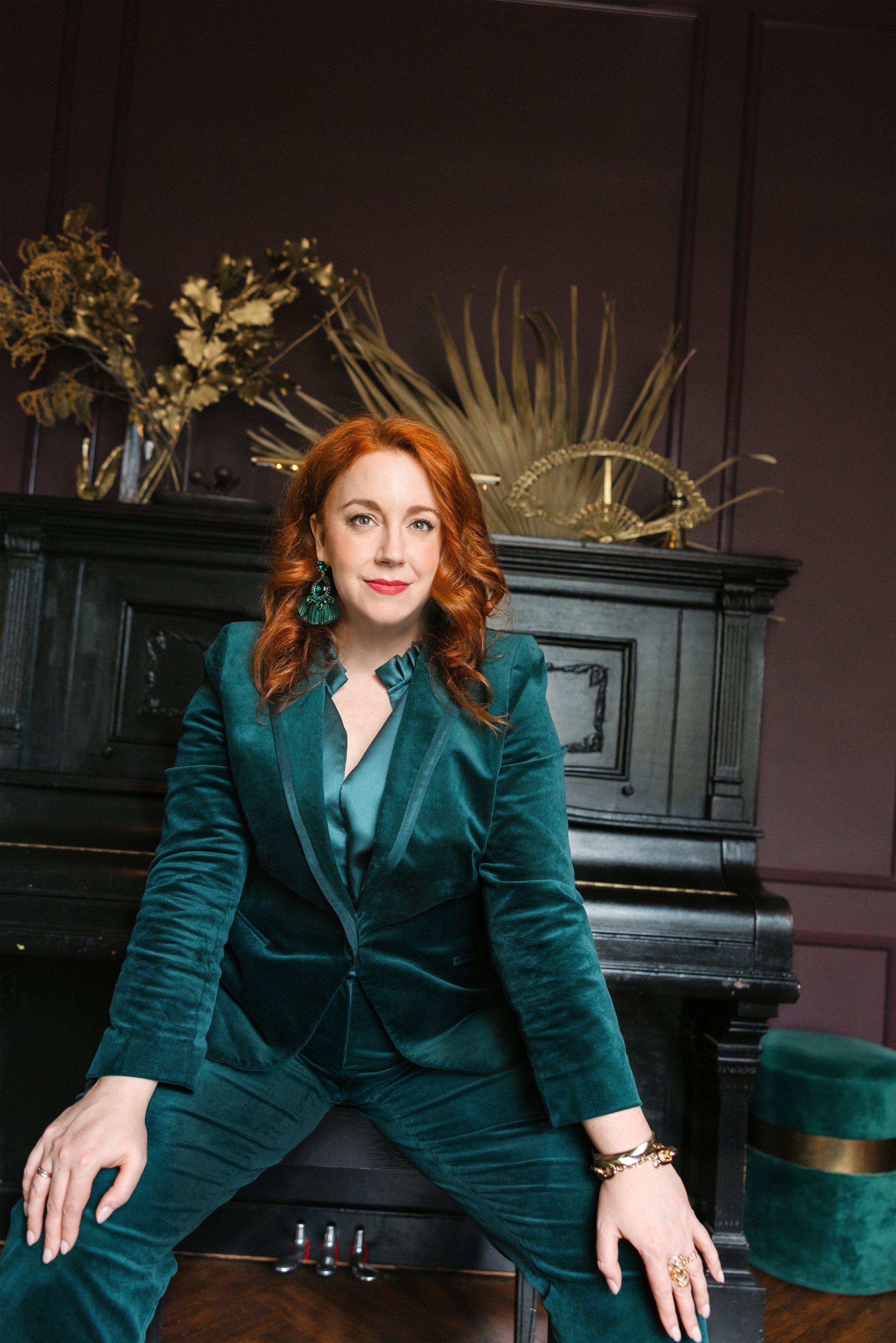 A woman with red hair wearing a teal velvet blazer and pants, sitting on a bench in front of a black piano with abstract gold and dark plant decorations on top, in a room with dark purple walls.