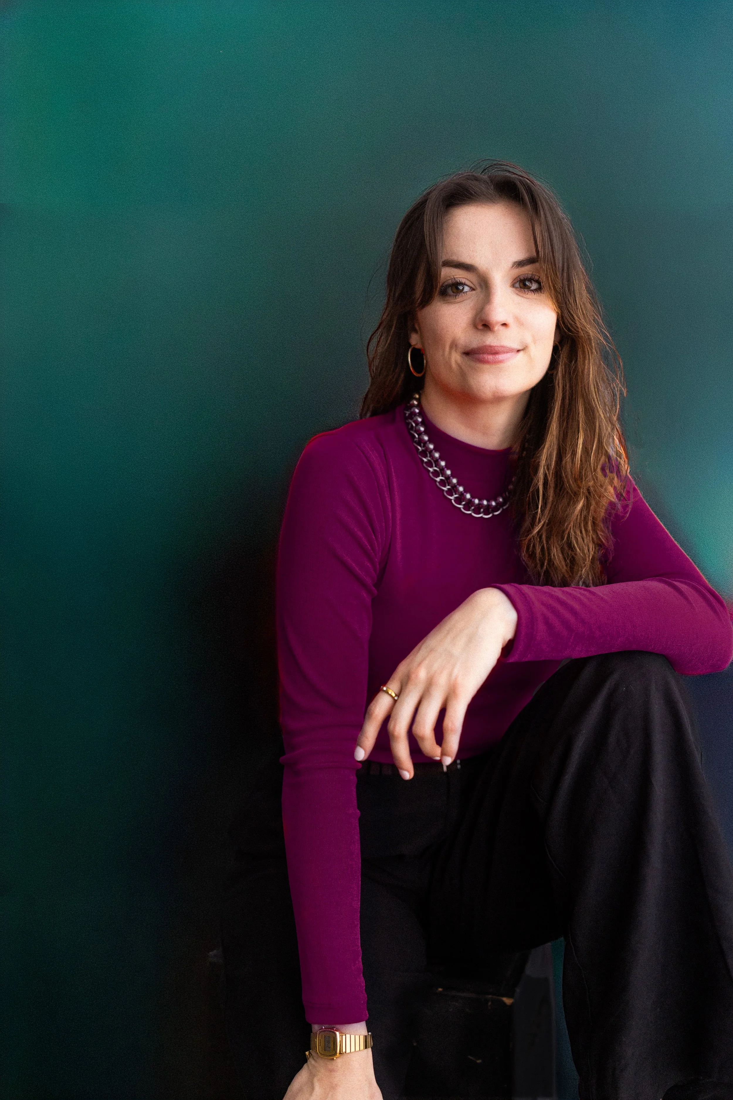 Claire Frances Sullivan, Right Hand Woman to the cofounders of Vital Voice Training, sitting against a dark green wall, wearing a purple long-sleeve top. She has dark hair, a warm smile, and an air of “I got you” competence.