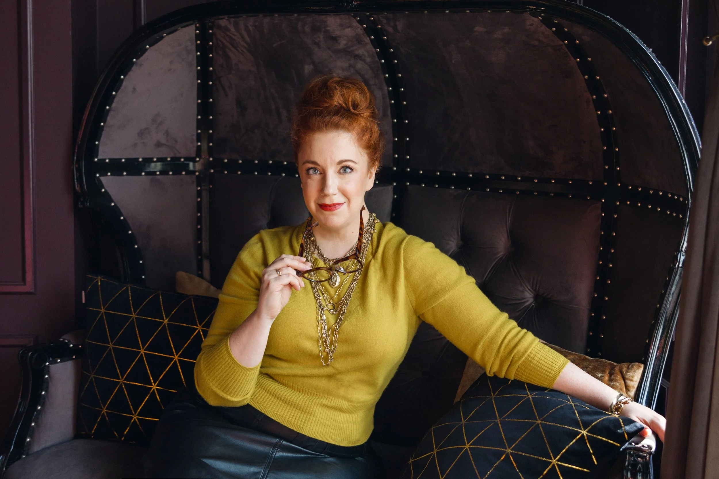 A woman with red hair sitting on a black, high-backed, tufted chair with decorative pillows, wearing a yellow sweater and holding eyeglasses, in a dimly lit room with dark furniture and walls.