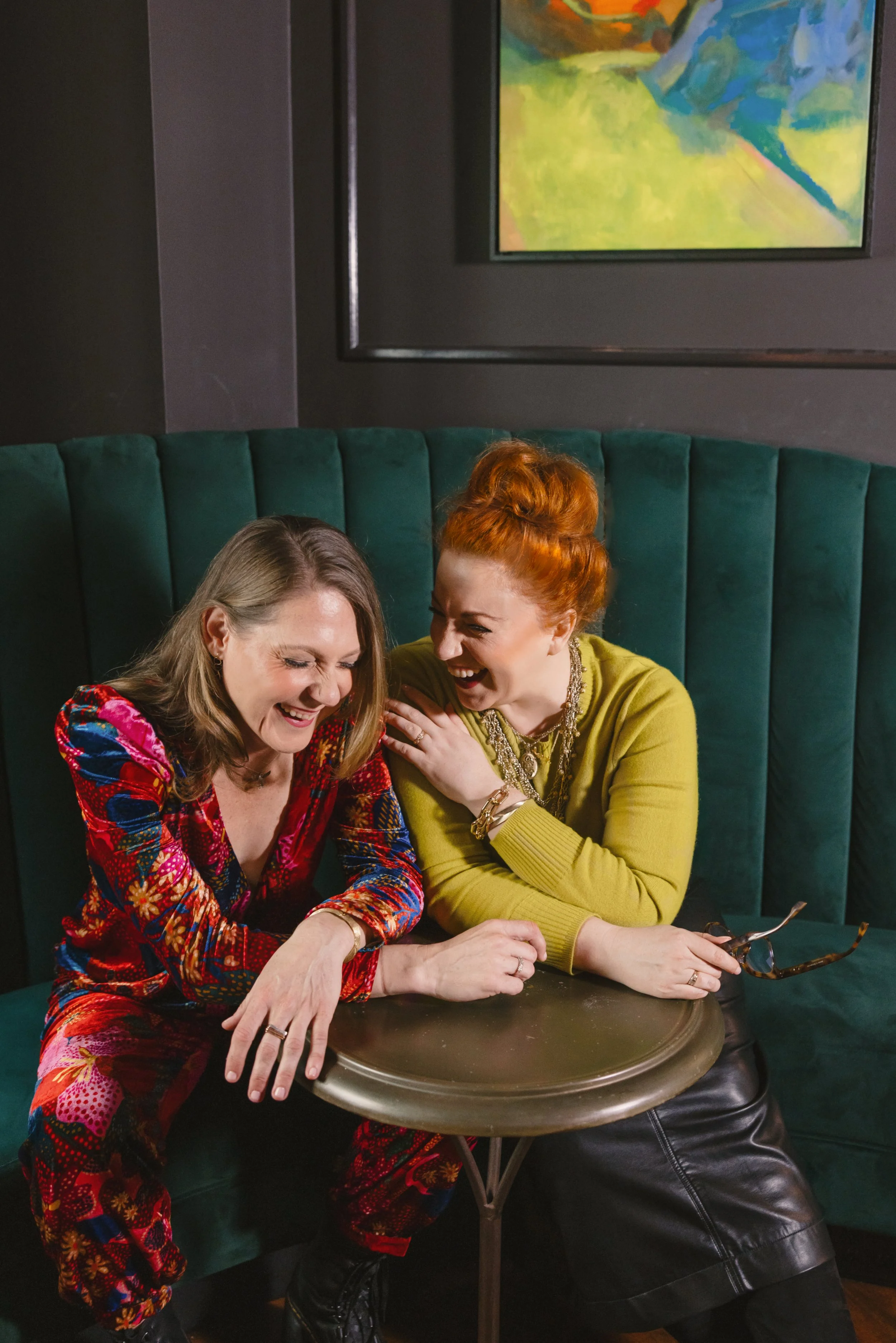 Two women sitting on a green velvet booth, laughing and sharing a joyful moment. One is wearing a colorful, patterned outfit, and the other is dressed in a green sweater with jewelry. They are leaning on a small round table in a cozy, dark-colored room with a framed painting on the wall behind them.