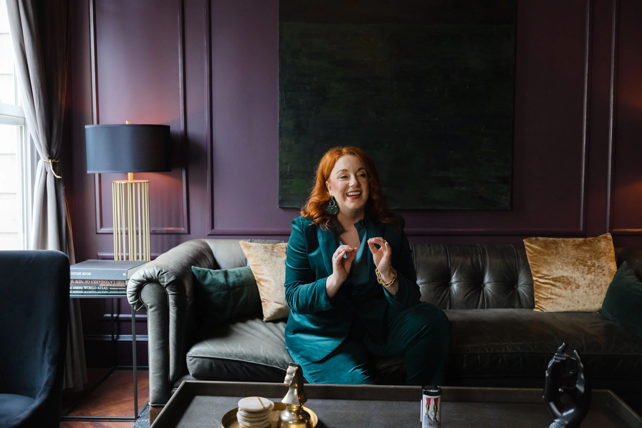 A woman with red hair, wearing a teal blazer and earrings, sitting on a dark leather sofa in a room with dark purple walls, a large black painting, and a window with curtains. She is smiling and gesturing with her hands.
