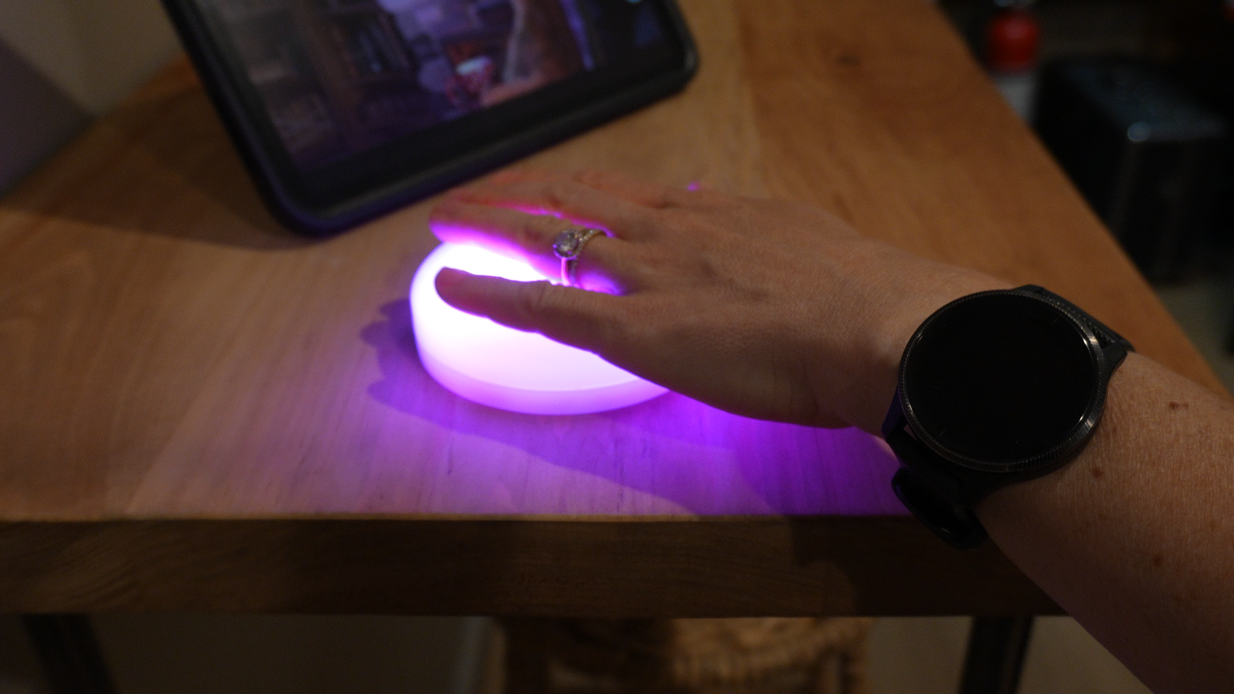 A person's hand with a ring and wristwatch is pressing on a glowing, purple lighted circle on a wooden surface, with a tablet nearby.
