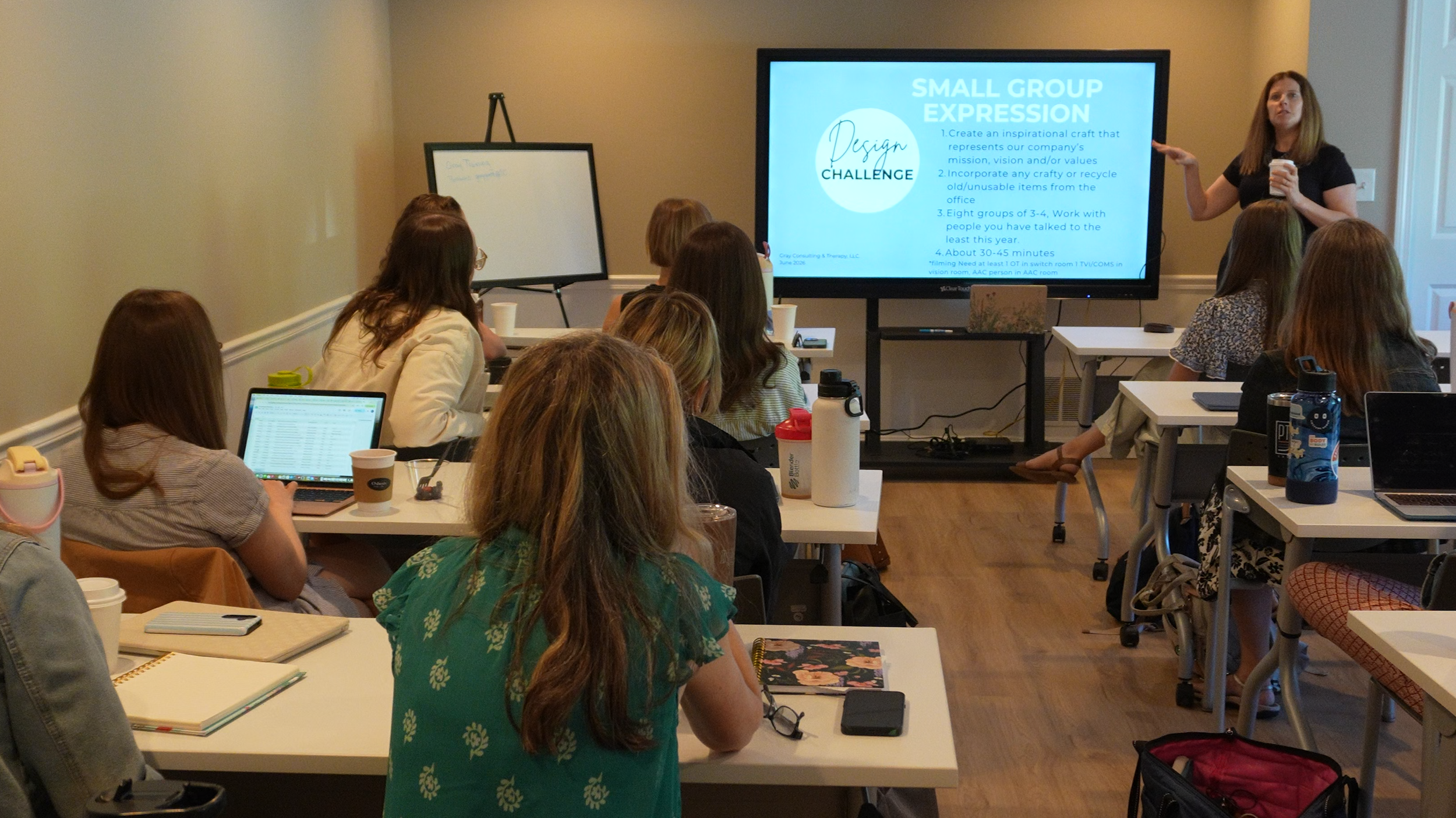 A group of women attending a workshop or presentation in a conference room, sitting at tables with notebooks, laptops, and drinks, listening to a woman standing by a large screen displaying a presentation slide.