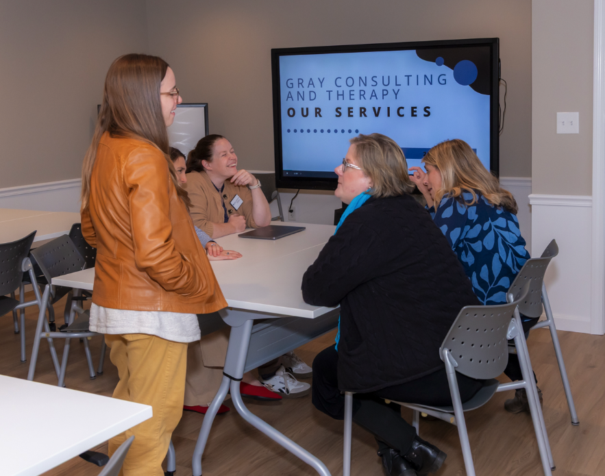 Group of people in a meeting room engaging in a discussion, with a woman standing and four women sitting around a table, a large screen displaying 'Gray Consulting and Therapy' in the background.