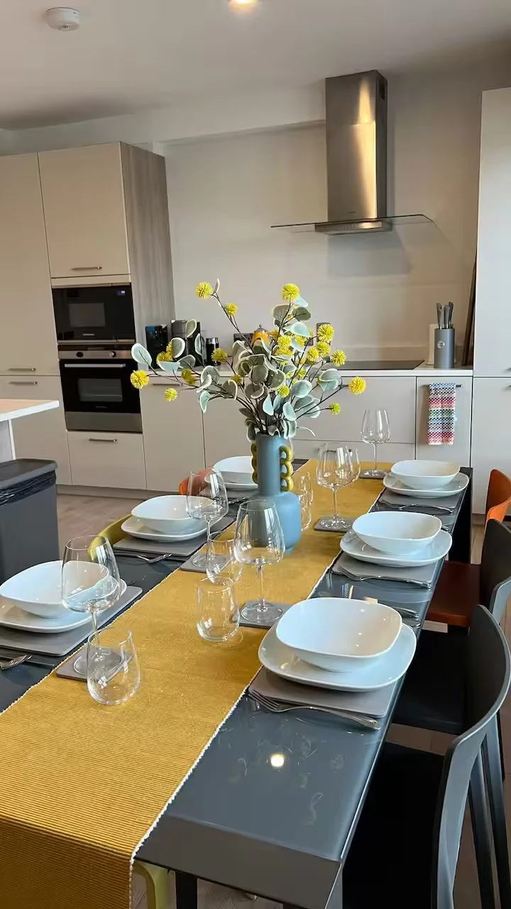 Dining table set for six with white bowls, wine glasses, and silverware, centered with a vase of yellow and green flowers. Kitchen in background with beige cabinets, a stove, and a stainless steel range hood.