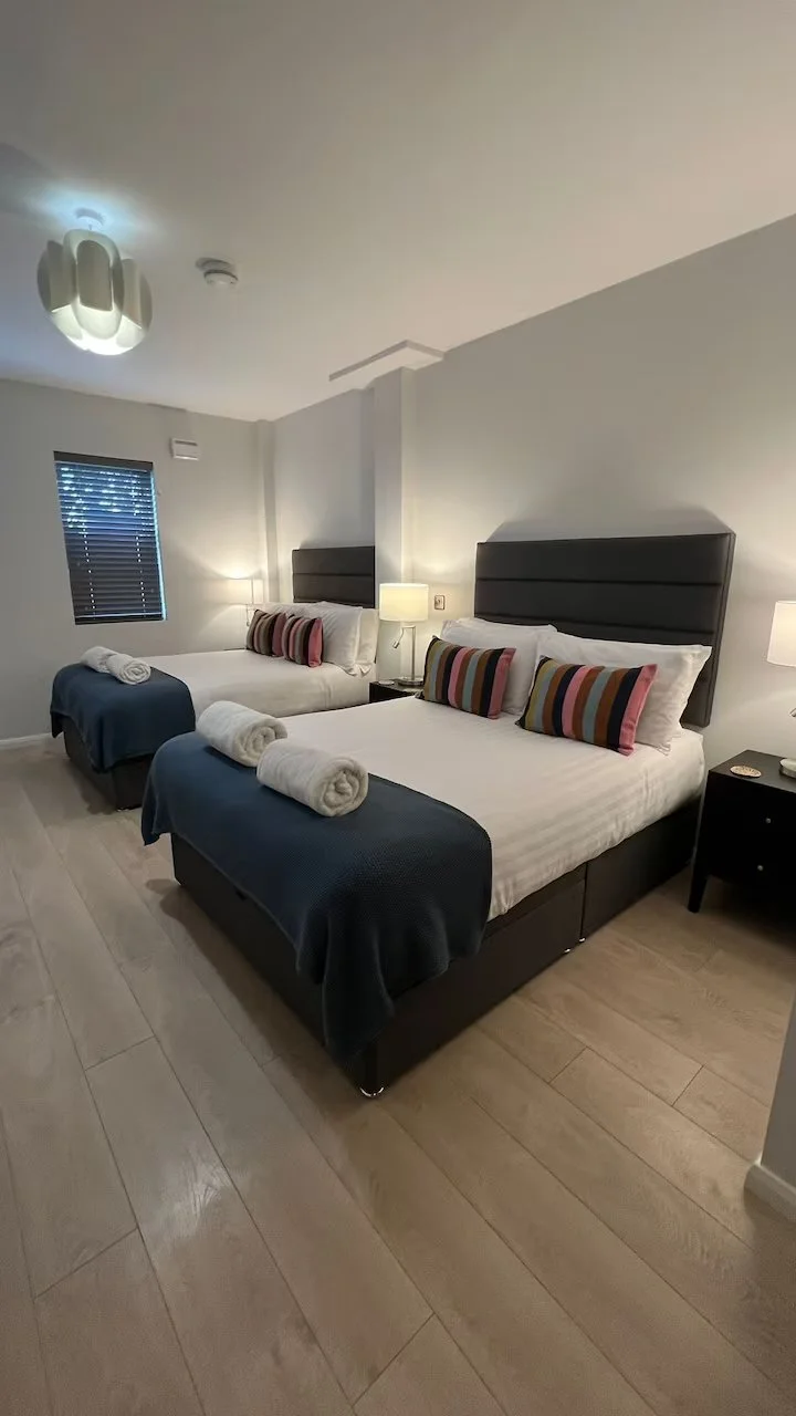 Two neatly made beds with dark headboards, each with striped pillows and white rolled towels at the foot, in a modern hotel room with light-colored wood flooring, bedside lamps, and a window with closed blinds.