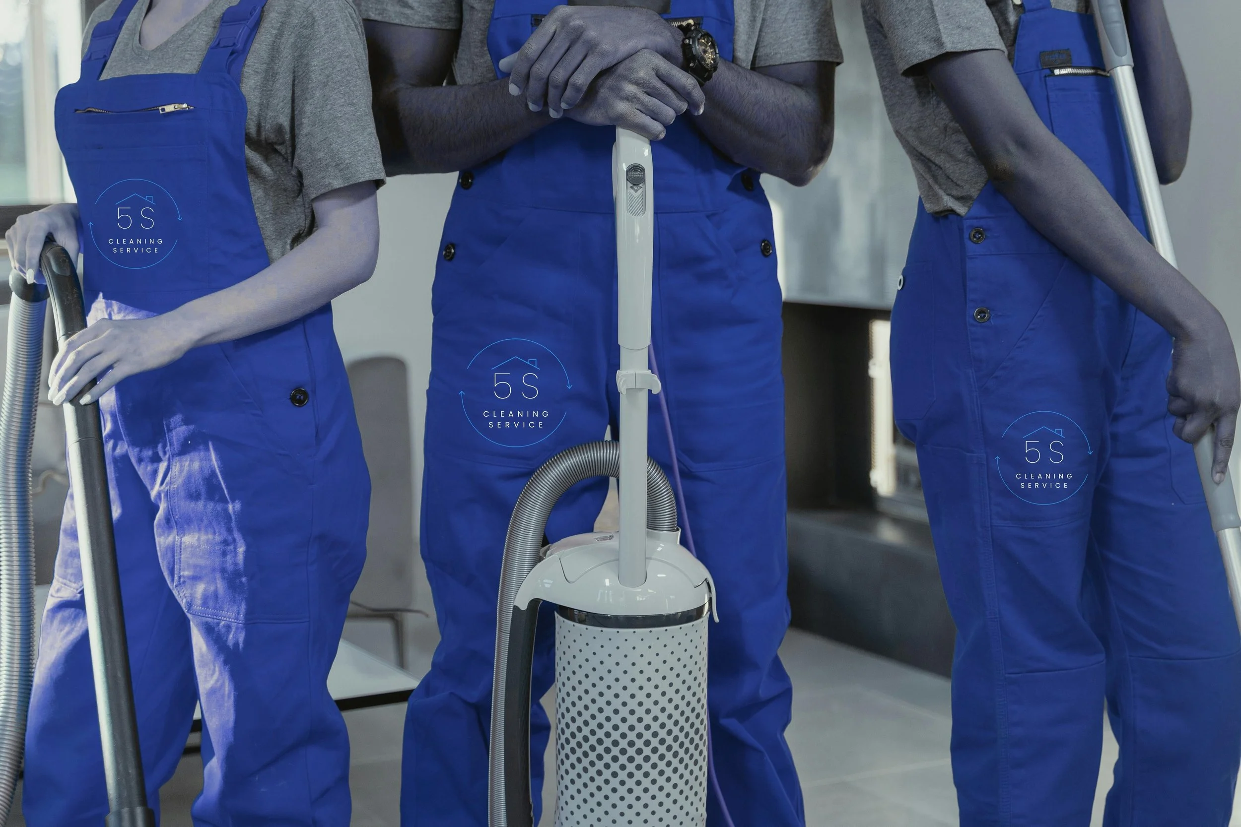 Three cleaning staff members wearing blue uniforms and gray shirts, holding vacuum cleaners, standing together in an indoor setting.