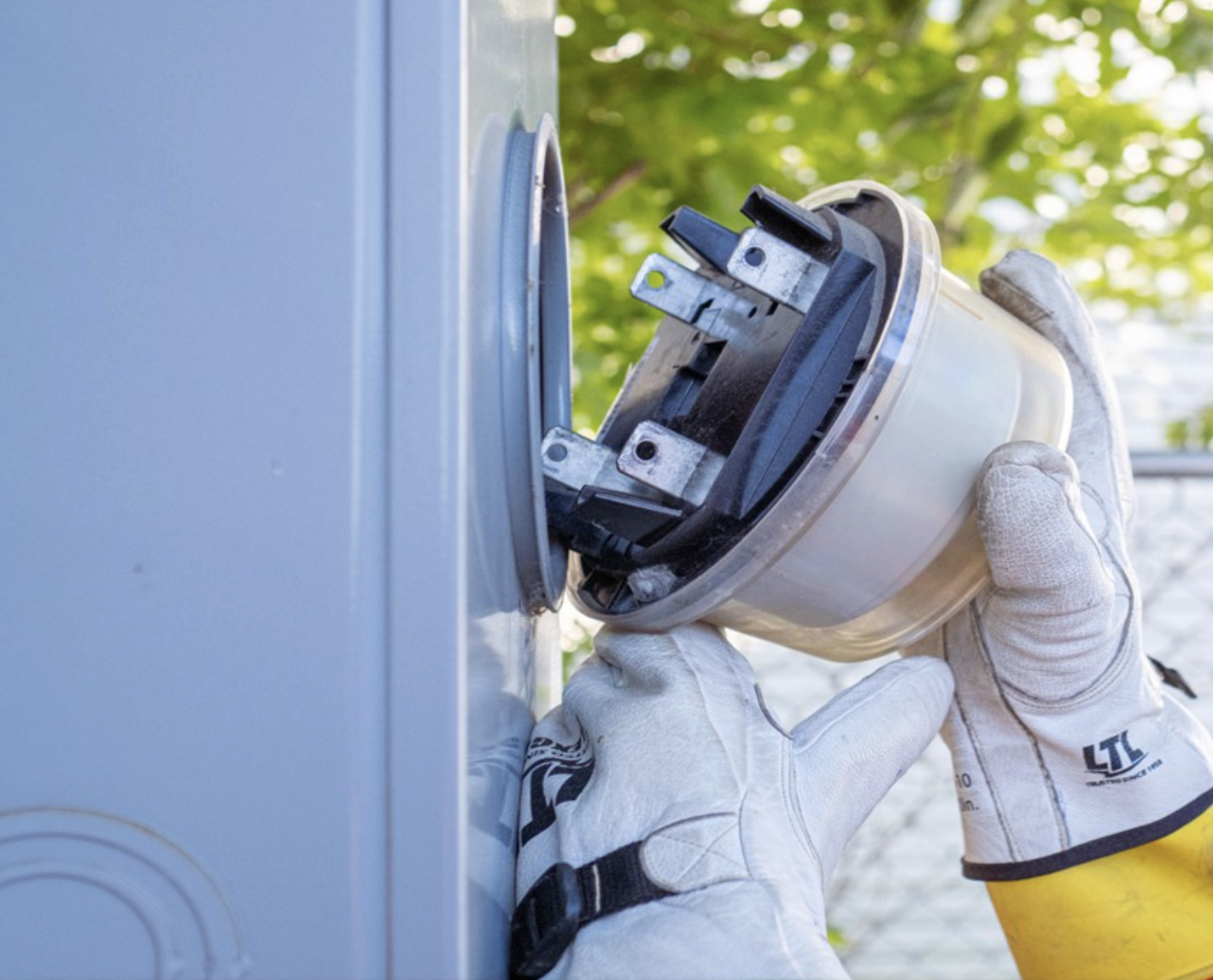 A person wearing white work gloves installs or repairs an electrical meter on an outdoor utility box, with trees and sunlight in the background.